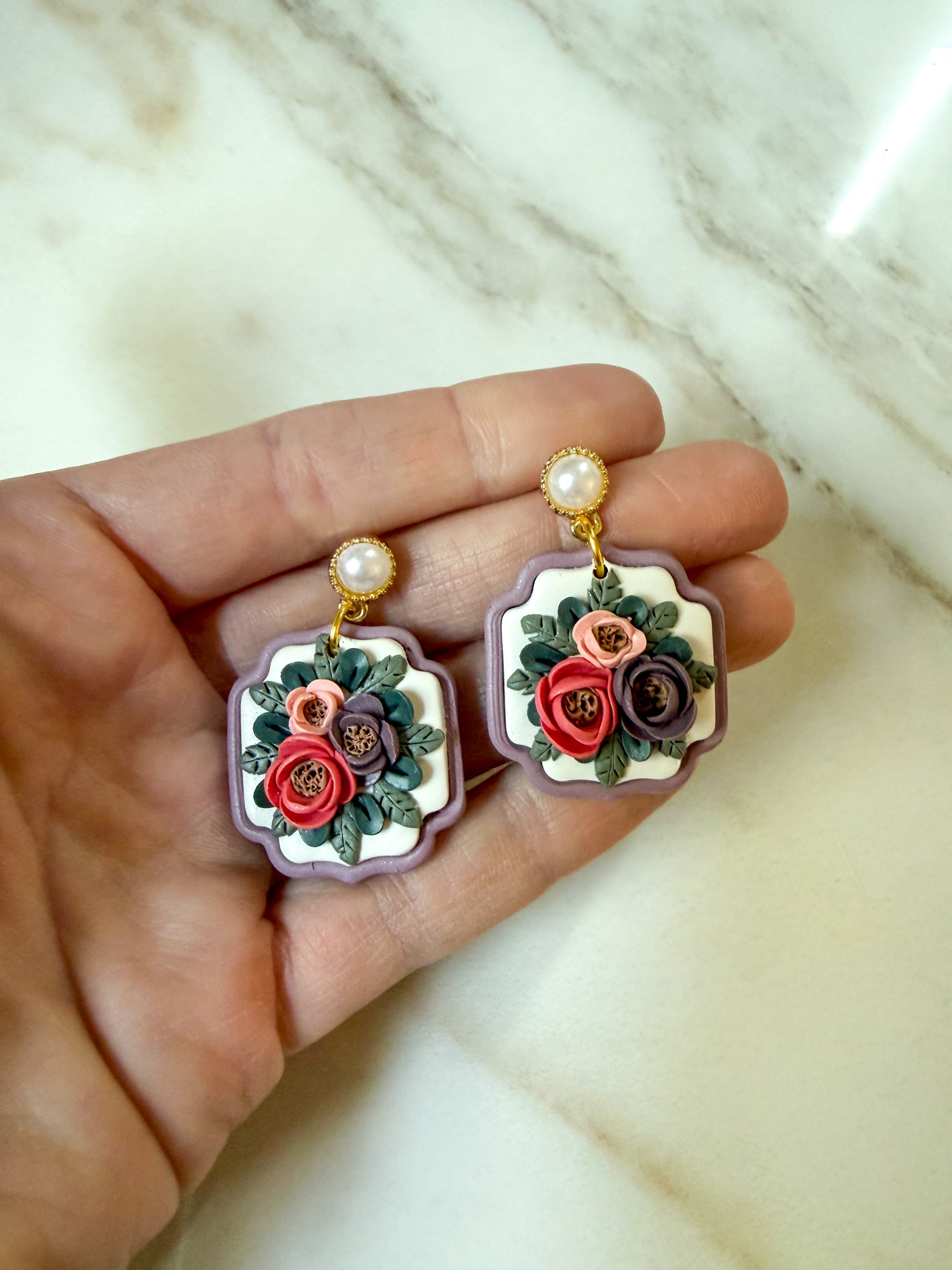 A pair of earrings held in a hand against a marble surface. They have hand rolled and individually placed petals. The posts of the earrings look like pearls. 