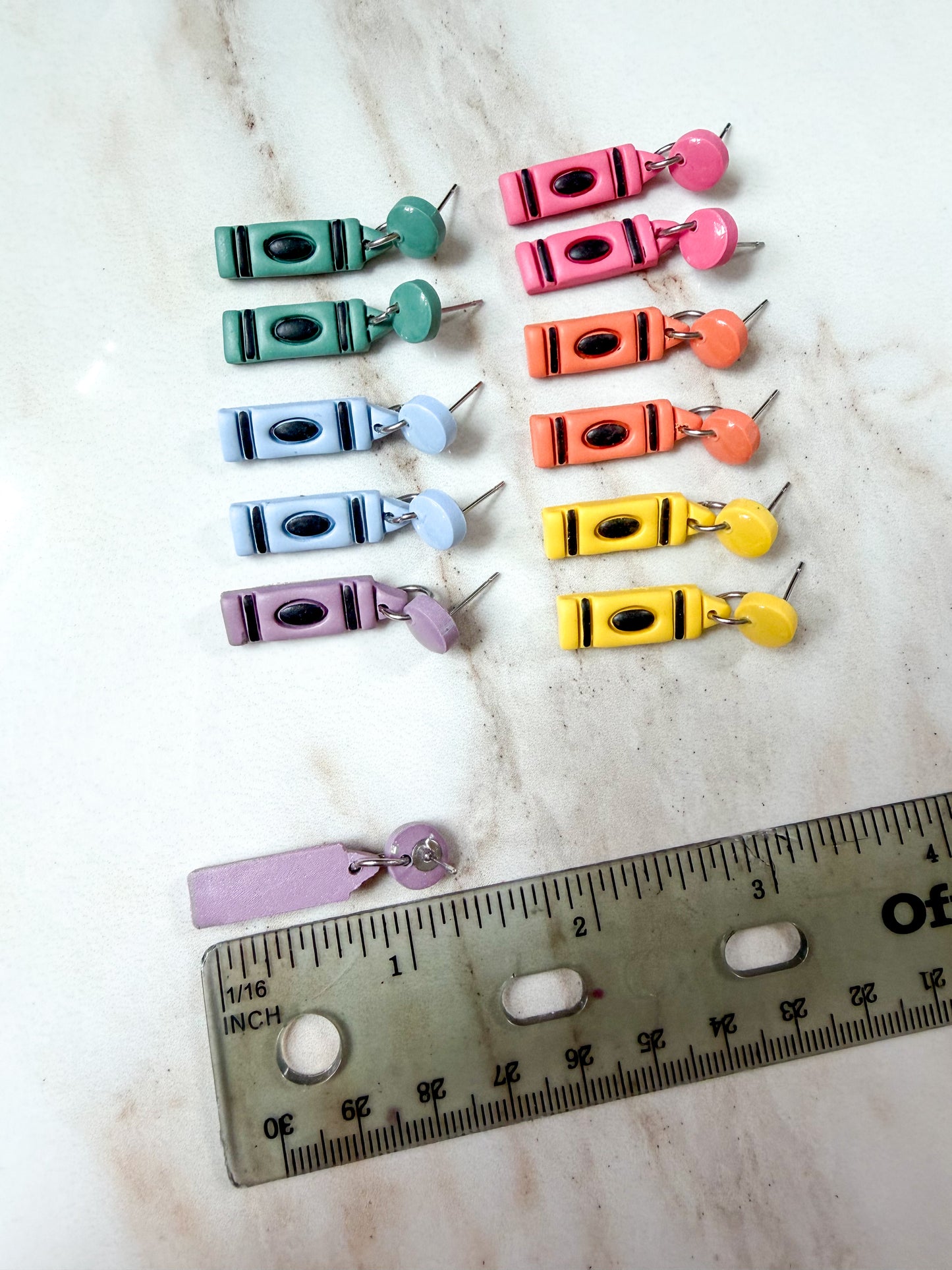 Twelve crayon-shaped stud earrings in assorted pastel and bright colors, arranged on a light marble surface with a metal ruler at the bottom for scale. About 1 1/4 inches long 