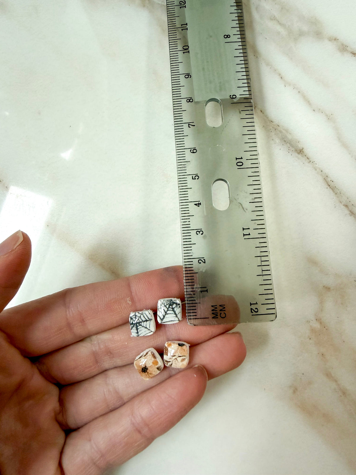 Small decorative square studs with Fall patterns are held in a person's hand next to a clear plastic ruler on a light marble surface. Less than 1cm long. 