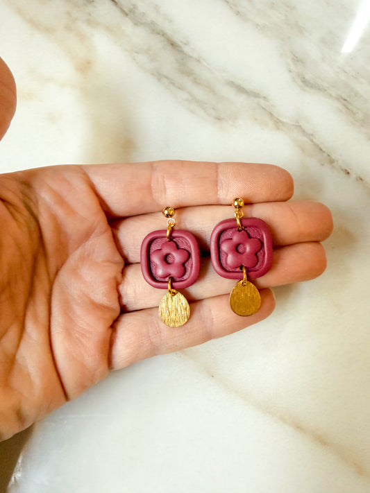 A pair of dangling earrings held in a person's hand. The earrings feature a burgundy square pendant with a flower motif, gold-tone posts, and small gold-tone round charms, set against a white marble background.