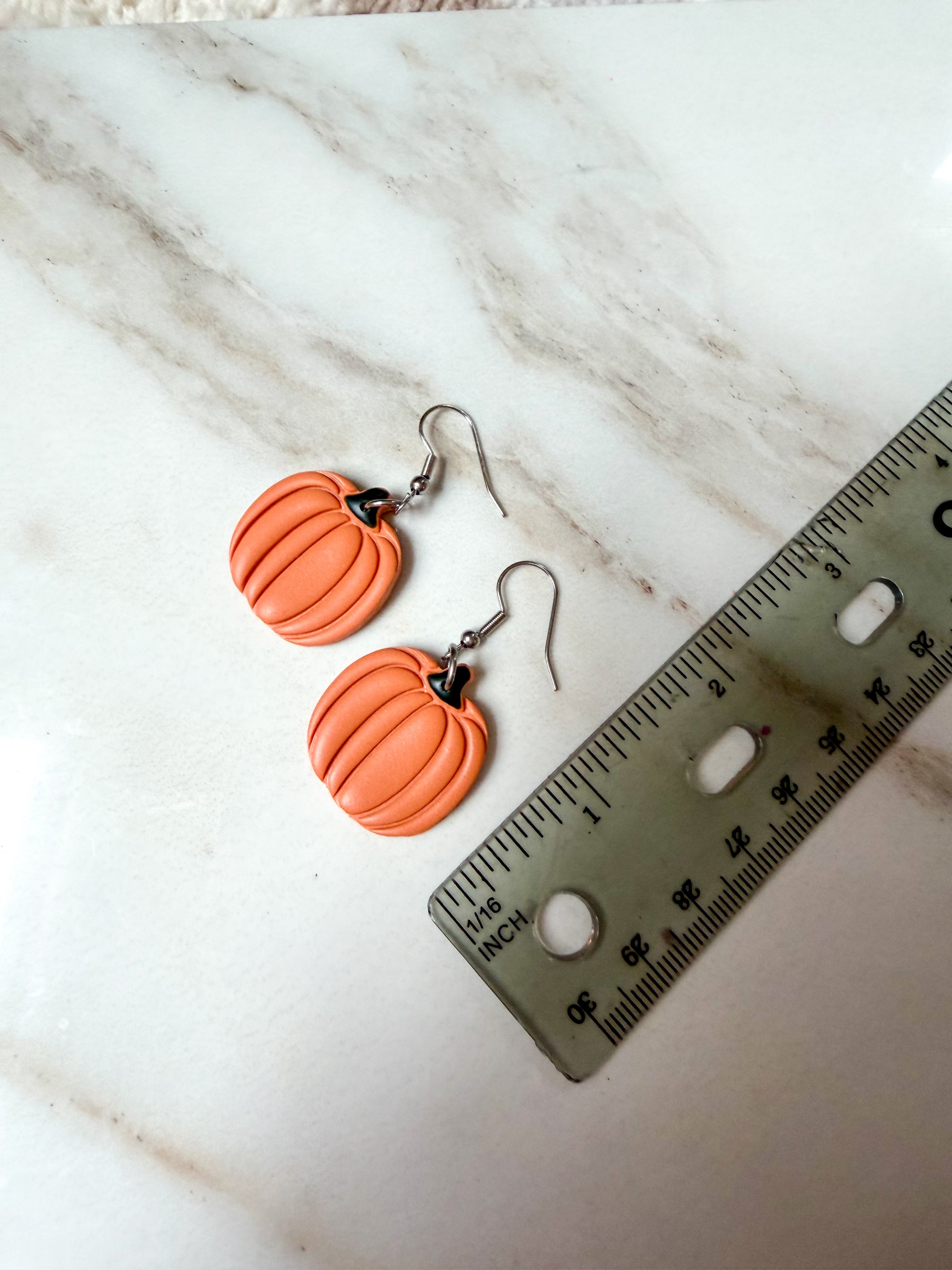 Orange pumpkin-shaped dangle earrings with silver fishhook wires, displayed on a white marble surface next to a ruler for scale. Around 2 inches in length. 
