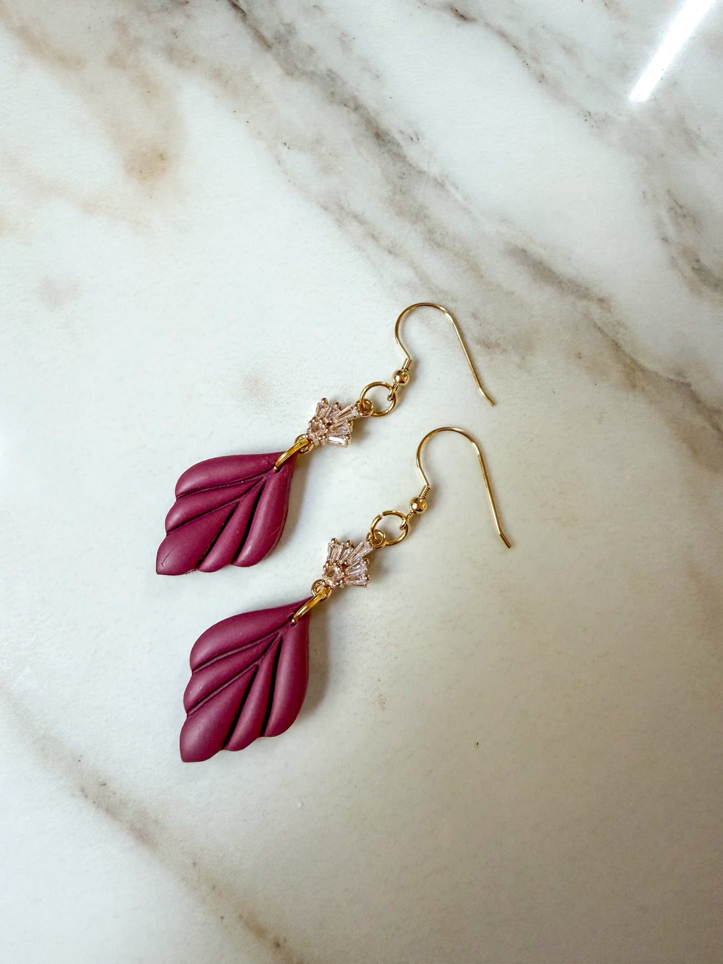 A pair of drop earrings with deep magenta leaf-shaped charms and gold hooks, resting on a white marble surface.