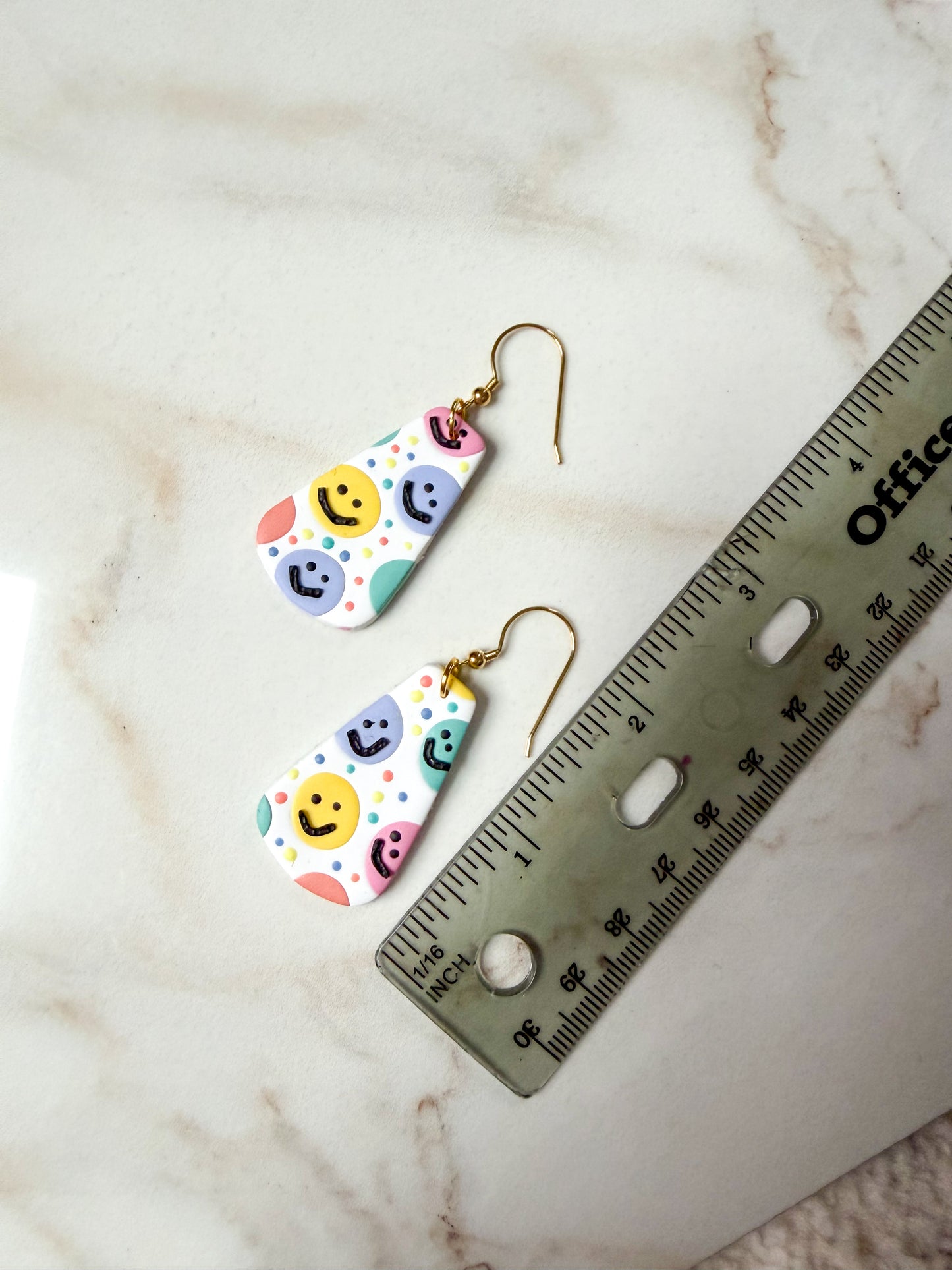 Colorful smiley face earrings on a marble surface with a ruler for scale. About 2 inches long.