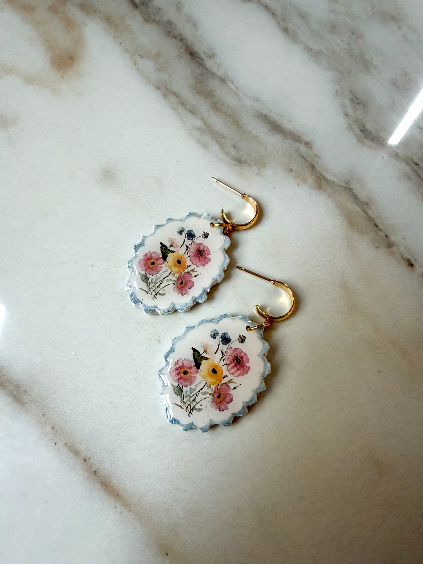 A pair of oval drop earrings with scalloped edges, featuring a floral design in pink, yellow, and blue on a white background, hanging from gold hooks on a white marble surface.