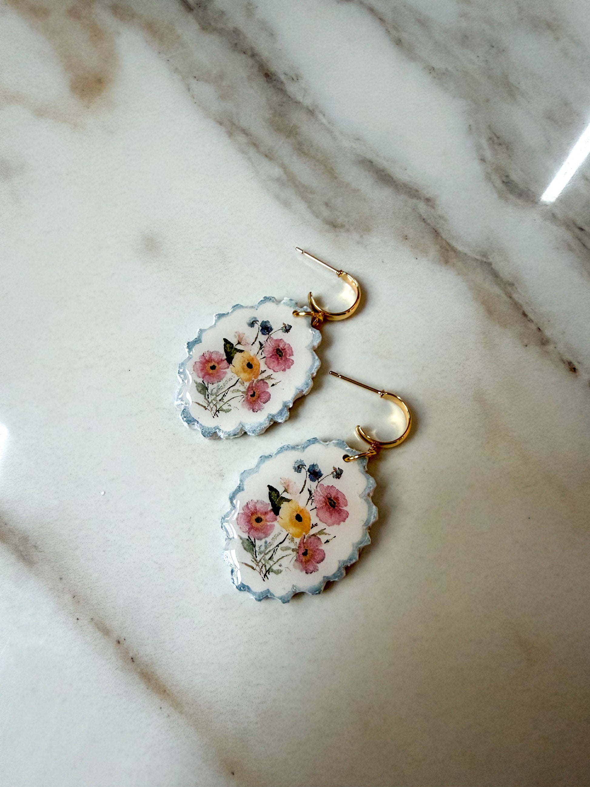 A pair of oval drop earrings with scalloped edges, featuring a floral design in pink, yellow, and blue on a white background, hanging from gold hooks on a white marble surface.