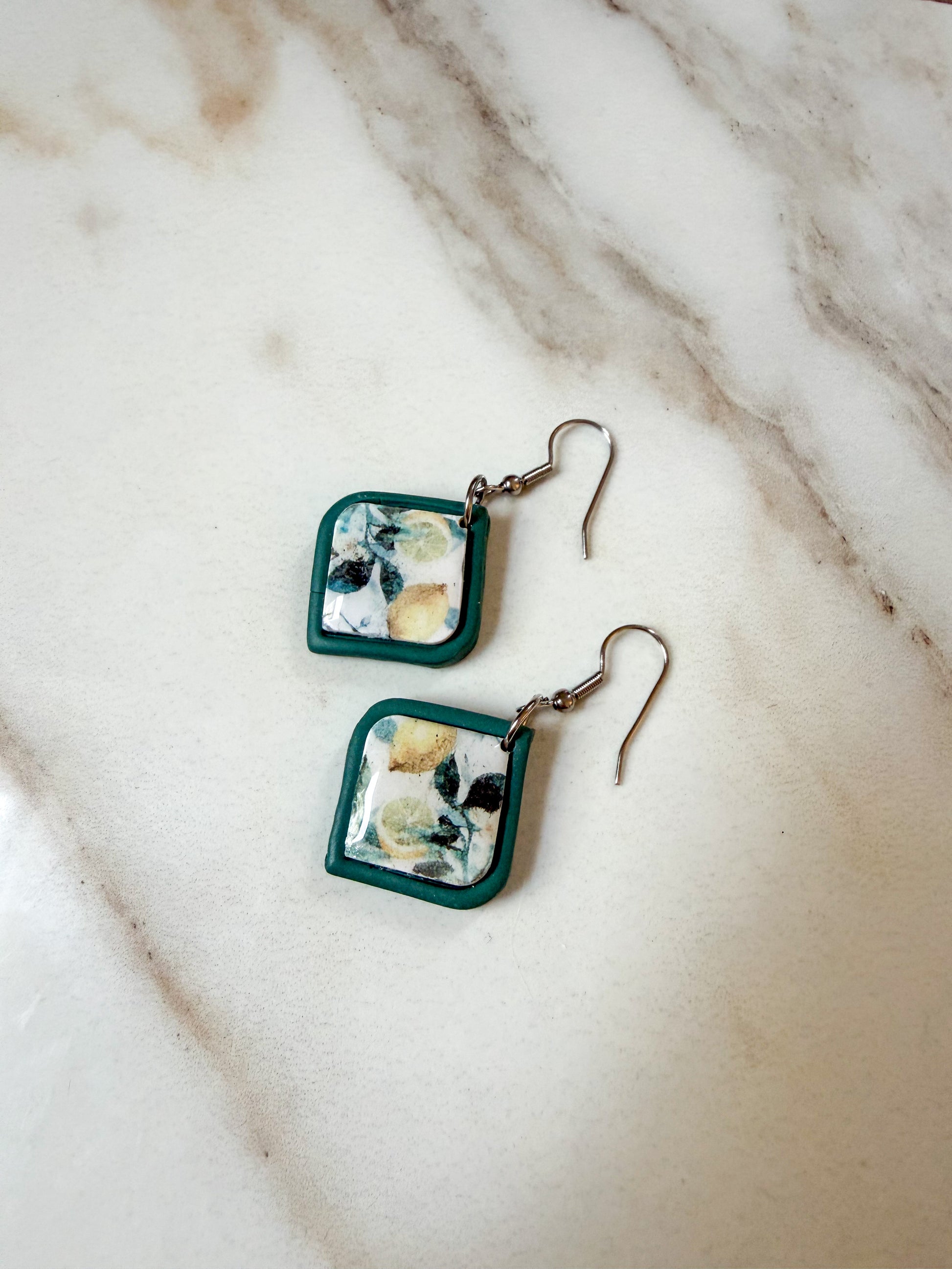 A pair of handmade square drop earrings with dark green borders and a white and lemon patterned center, hanging from silver fishhook wires on a light marble surface.