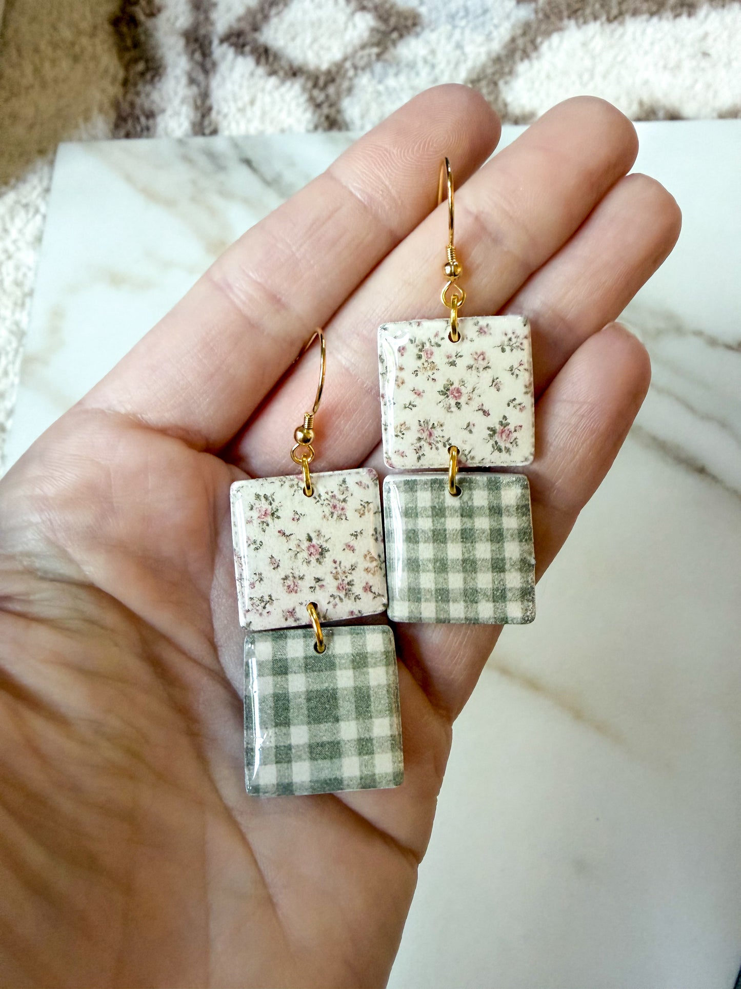A hand holding a pair of square dangle earrings with gold fishhook wires. The earrings feature two stacked patterned square charms: one with a small floral print and one with a green-and-white gingham pattern.