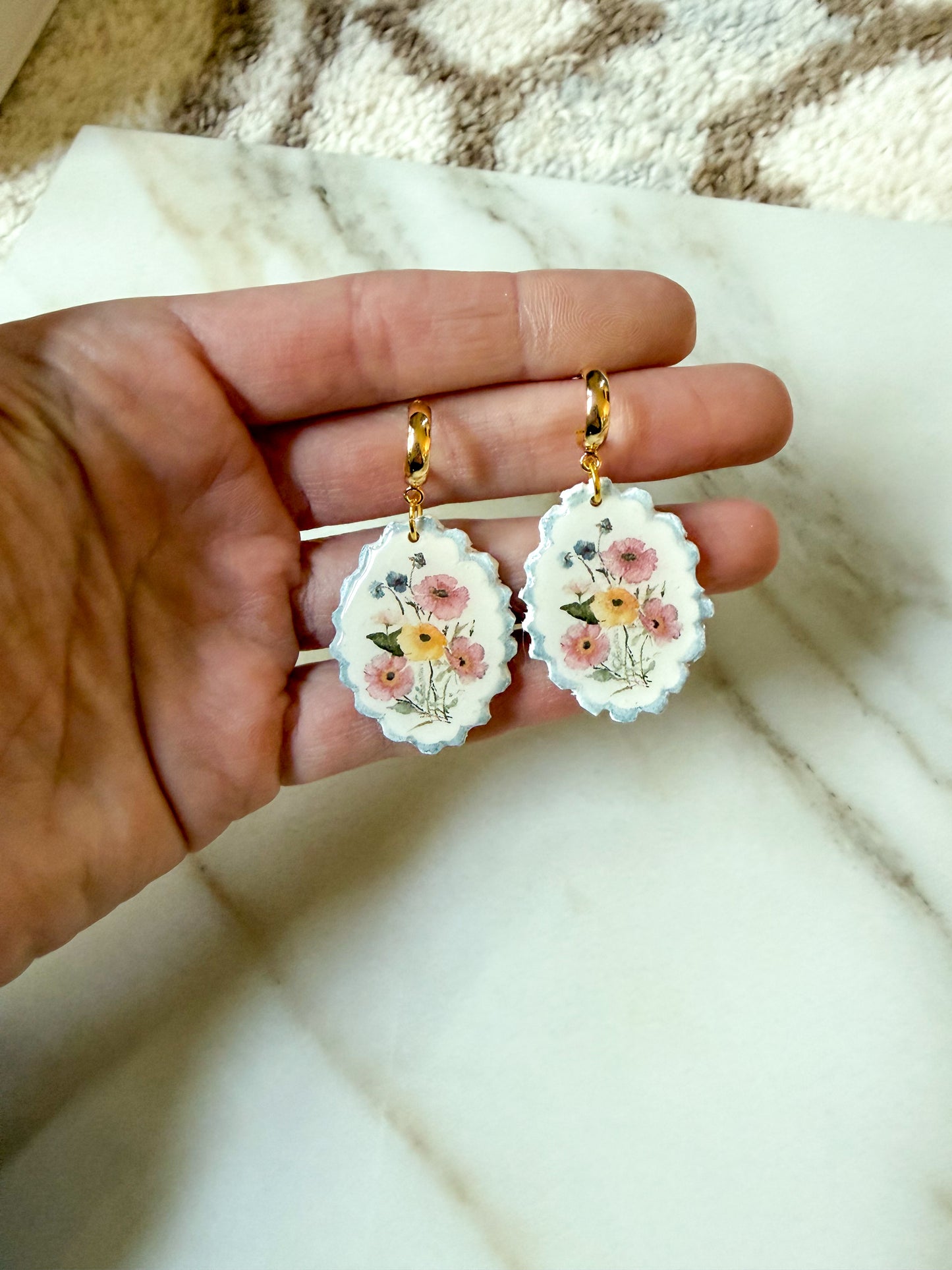 A hand holding a pair of white scalloped oval earrings with a delicate pink, yellow, and green floral design, suspended from gold-tone huggy posts, set against a white marble surface.