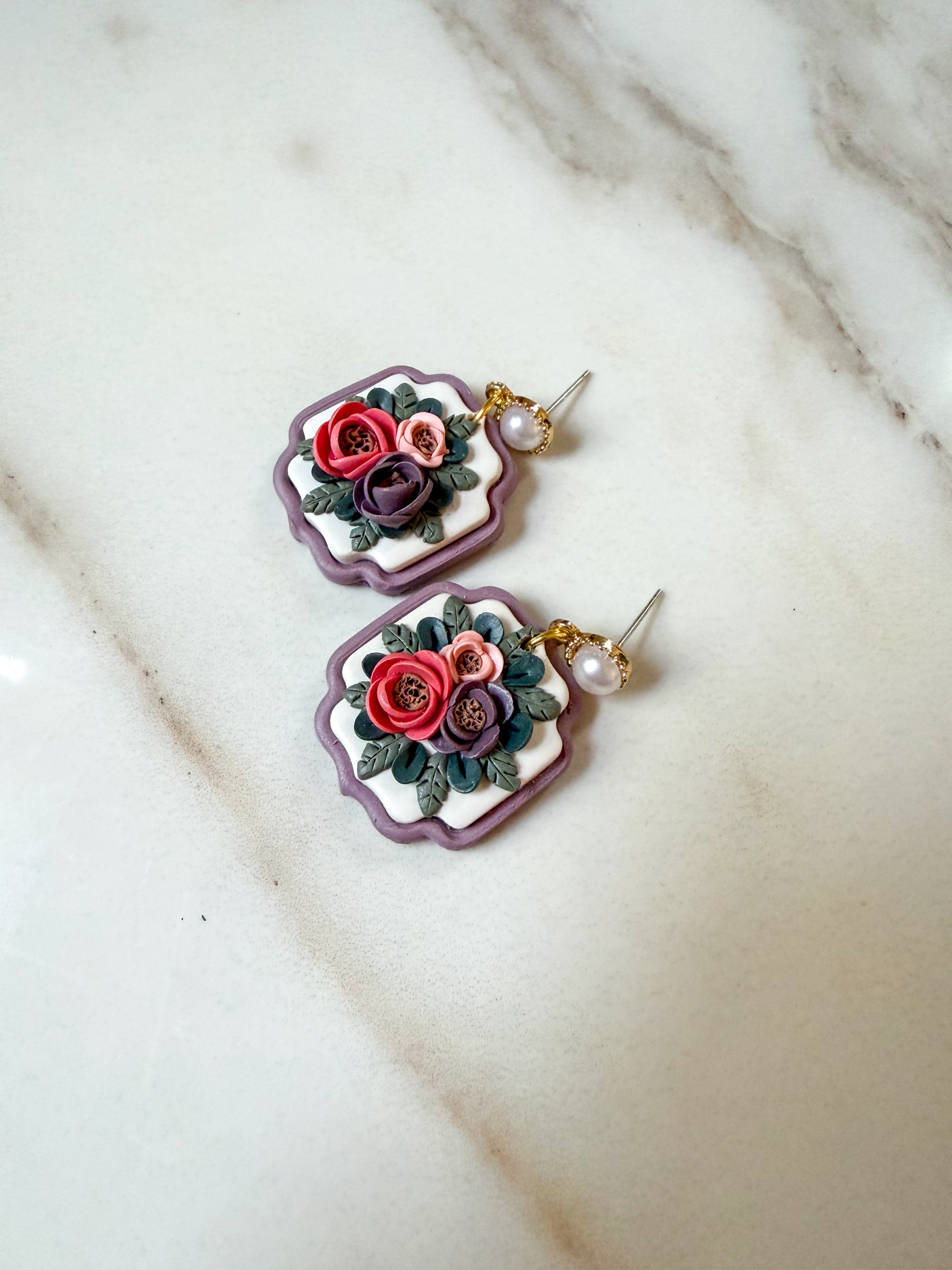 A pair of handmade polymer clay dangle earrings with floral designs, resting on a white marble surface. The earrings feature purple scalloped borders, white centers, and detailed red, pink, and purple flowers with green leaves, suspended from gold-toned posts with small pearl-like beads.