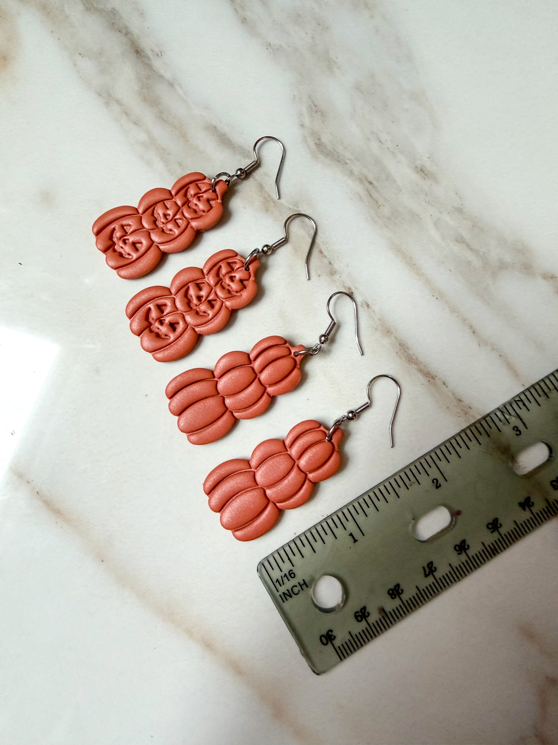 Four orange clay dangle earrings with silver fishhook wires, arranged on a white marble surface next to a ruler for scale. Around 2 inches in length. 