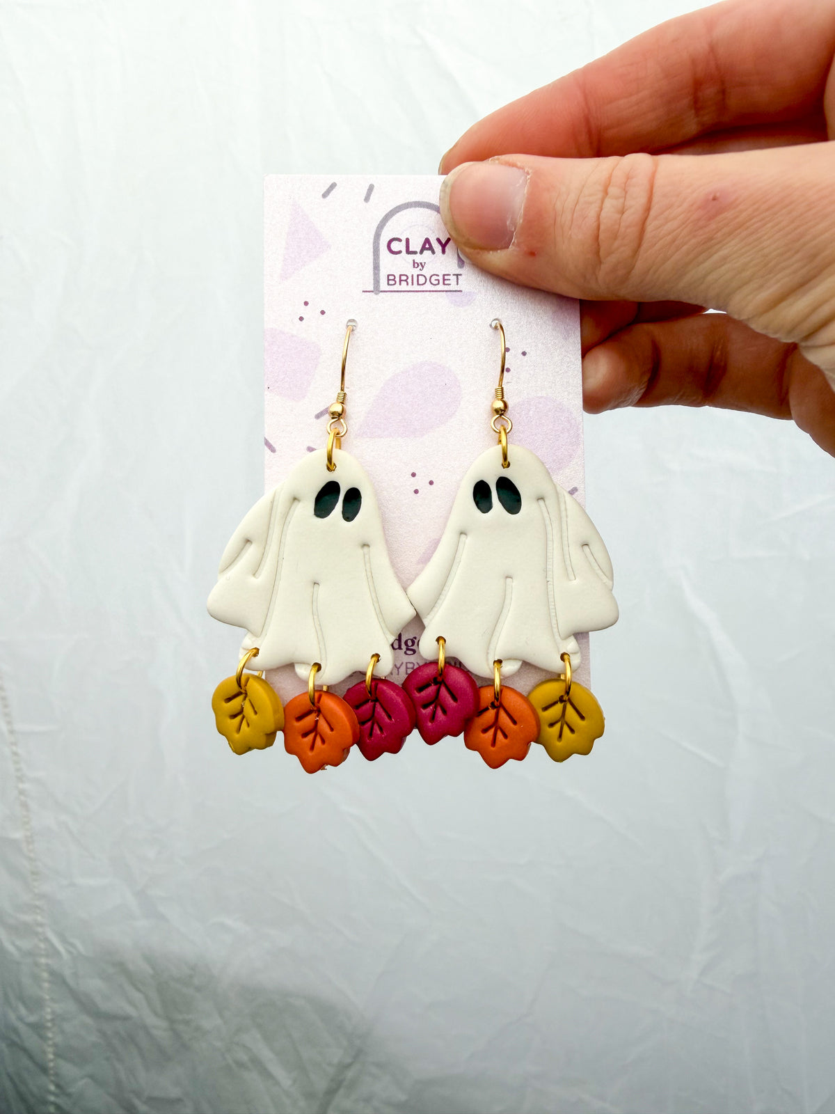 A pair of handmade polymer clay dangle earrings featuring white ghost shapes with black eyes and colorful autumn leaf charms in yellow, orange, and pink, displayed on a branded card held by a hand.