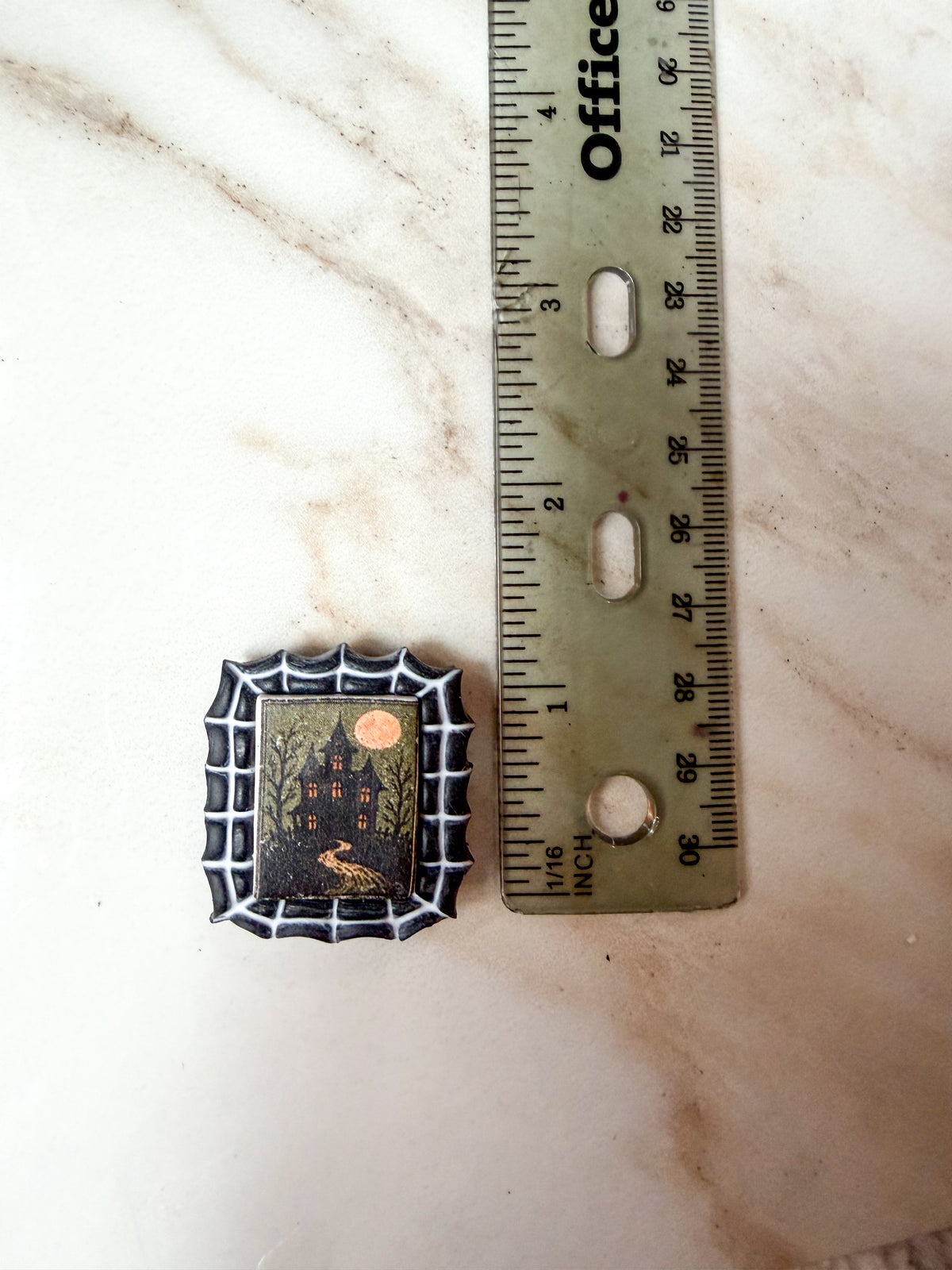 A small rectangular black spiderweb magnet with a haunted house illustration, shown next to a ruler on a light marble surface. A little over an inch long. 