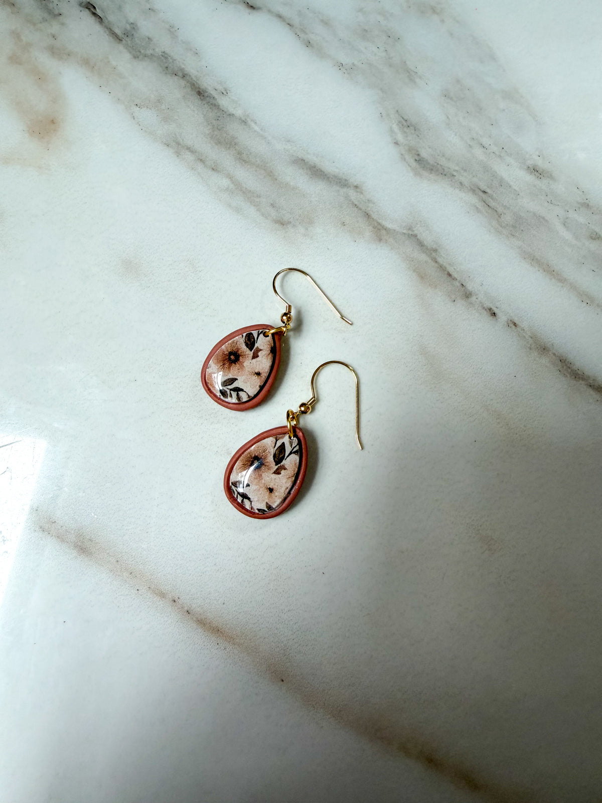 A pair of teardrop-shaped dangle earrings with gold hooks and reddish-brown borders, featuring a light floral pattern, displayed on a white marble surface.