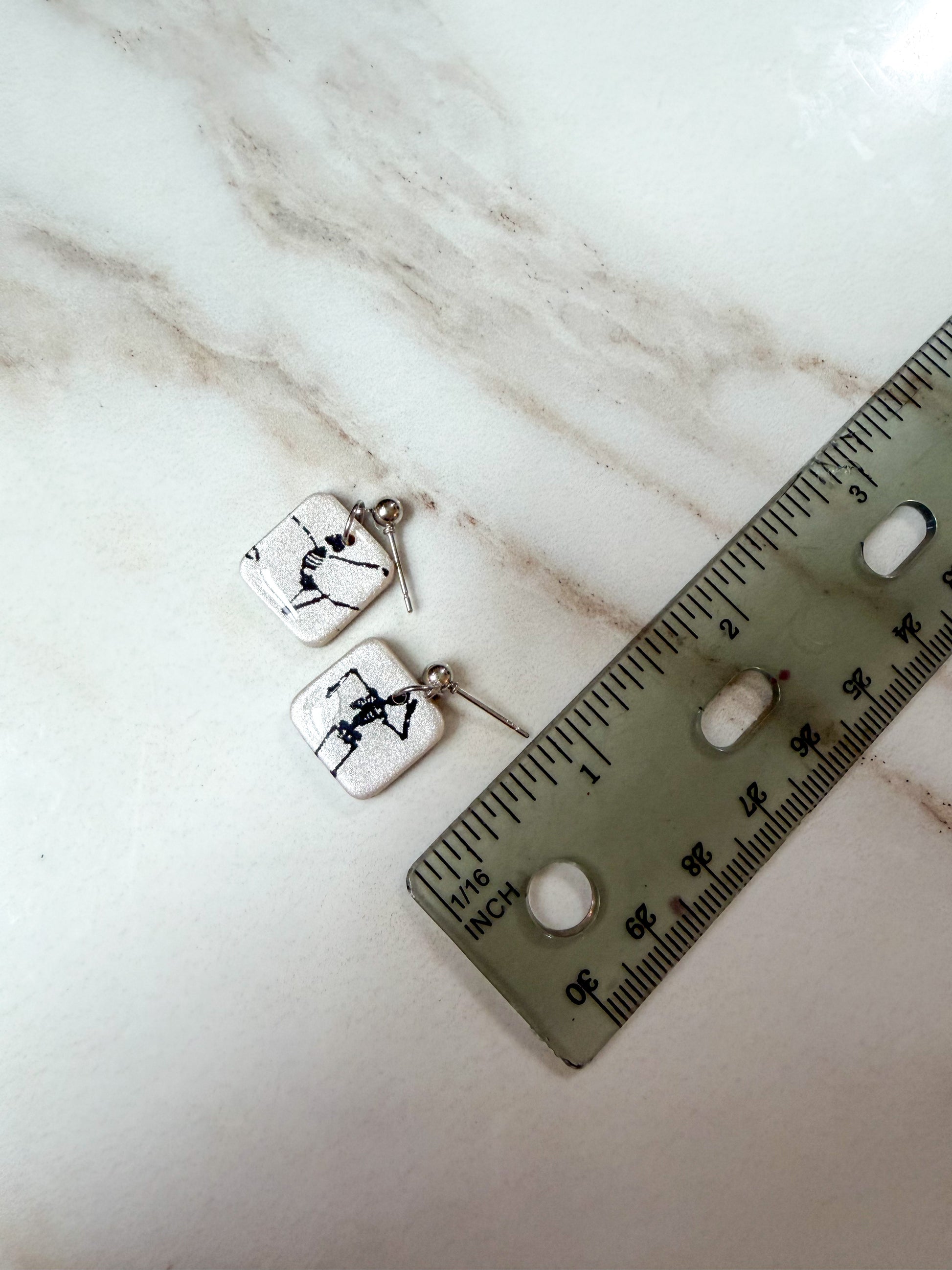 A pair of silver-toned square dangle earrings with black skeleton illustrations on a white marble background, shown next to a ruler on a light marble surface. 3/4 inches in length 