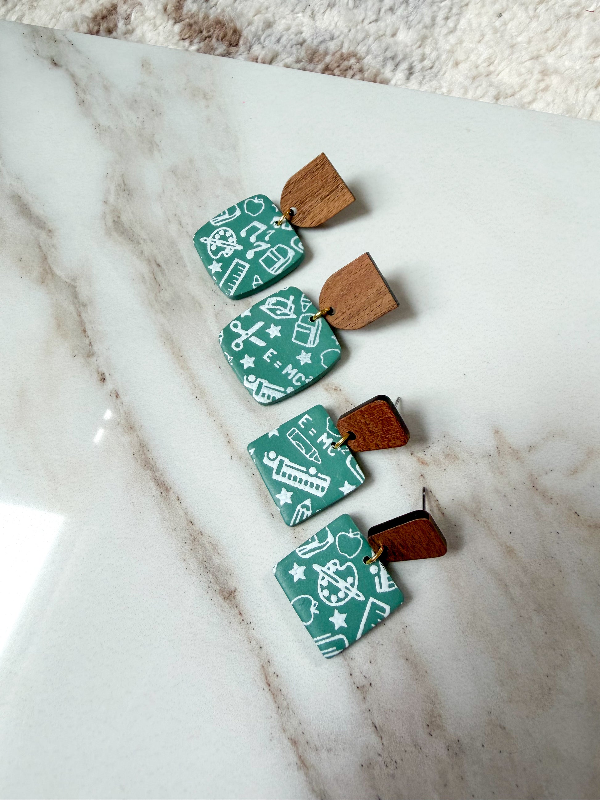 Four earrings on a marble surface. They are green to resemble old chalkboards, with white school-themed scribble designs. The posts are wood. 