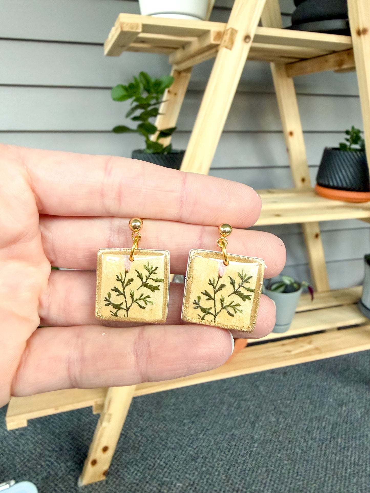 Gold square earrings with green leaf design held by a hand, with a wooden shelf and plants in the background.