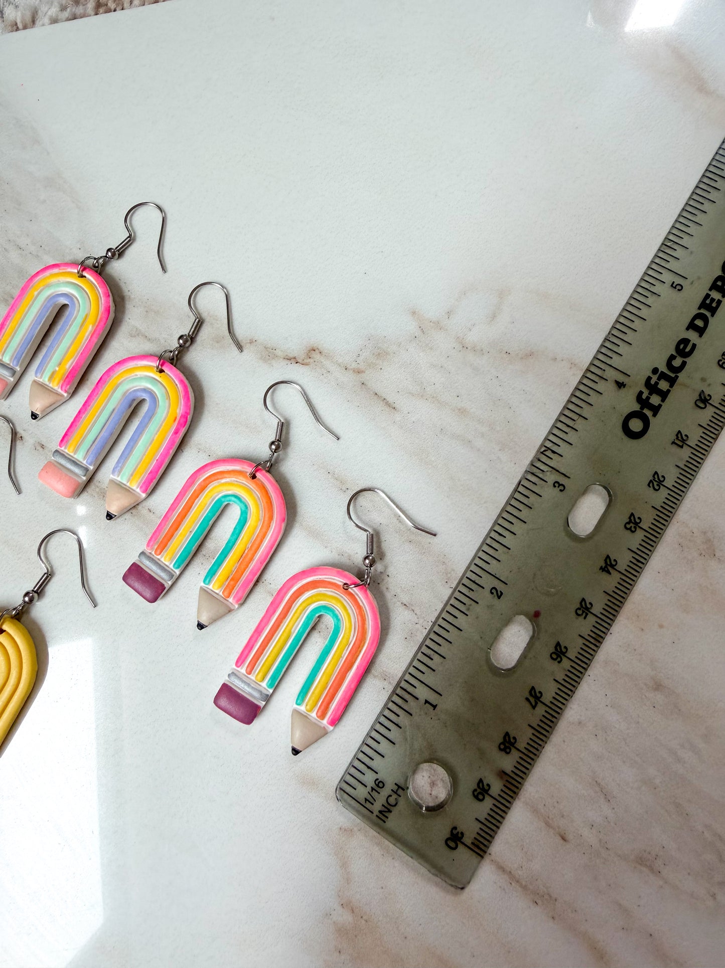 A set of handmade pencil-earrings with rainbow-colored bodies and silver fishhook wires, displayed on a white marble surface next to a metal ruler for scale. About 2 inches long. 