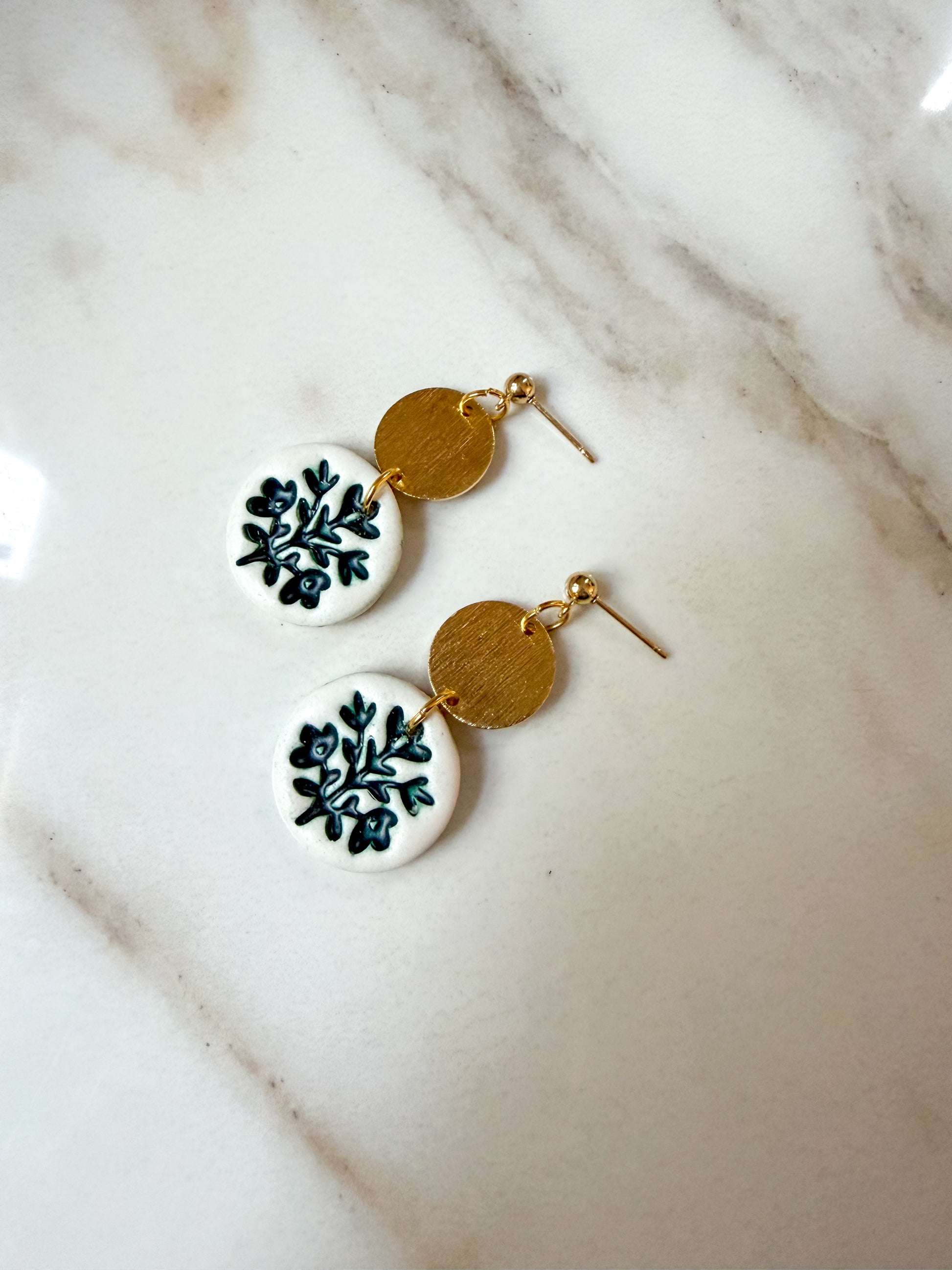 A pair of handmade dangle earrings with white circular discs featuring a green floral pattern, connected by textured gold discs to gold French hook ear wires, displayed on a white marble surface.
