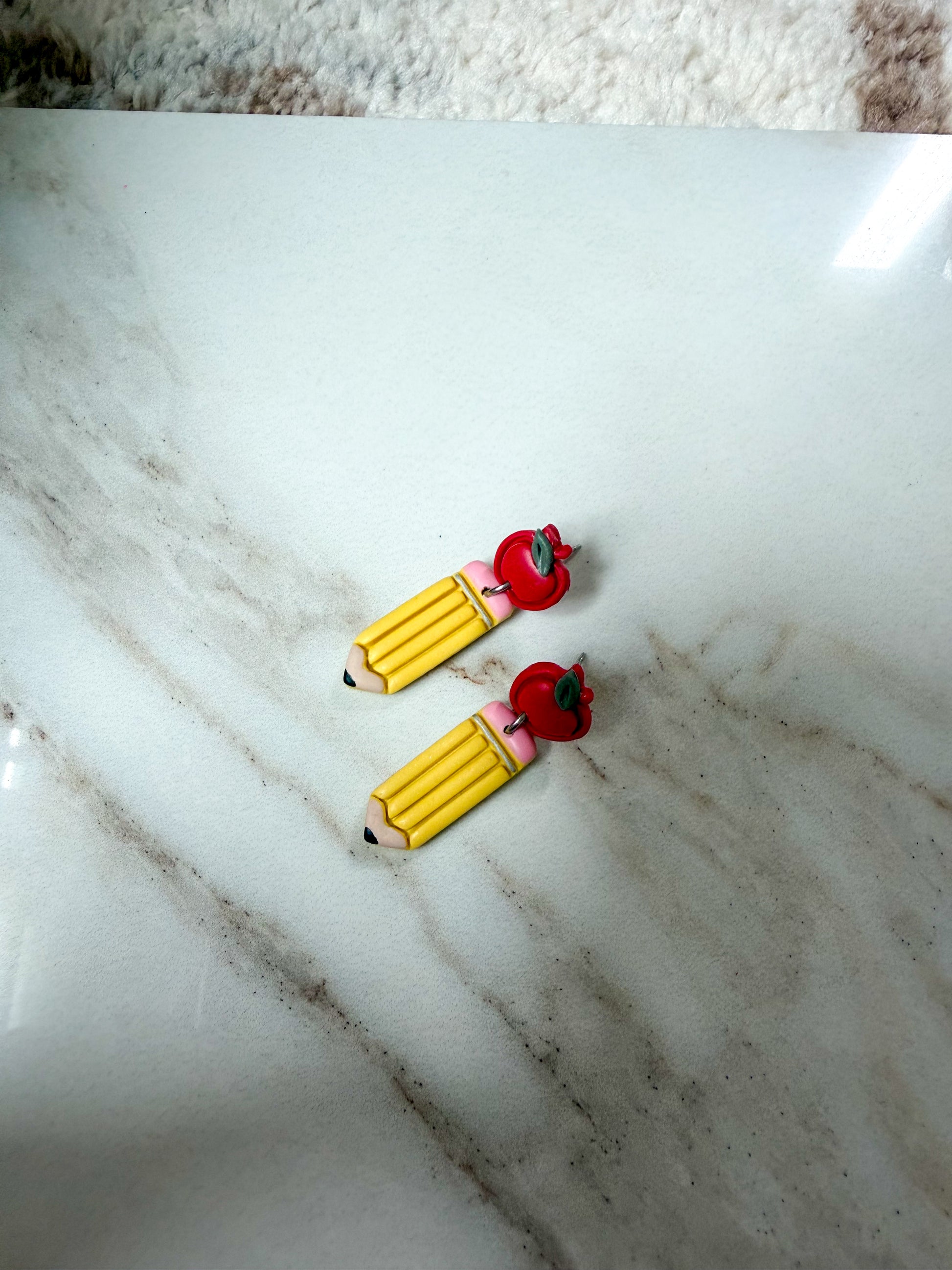 Two small pencil-shaped accessories with red apple tops resting on a white marble surface.