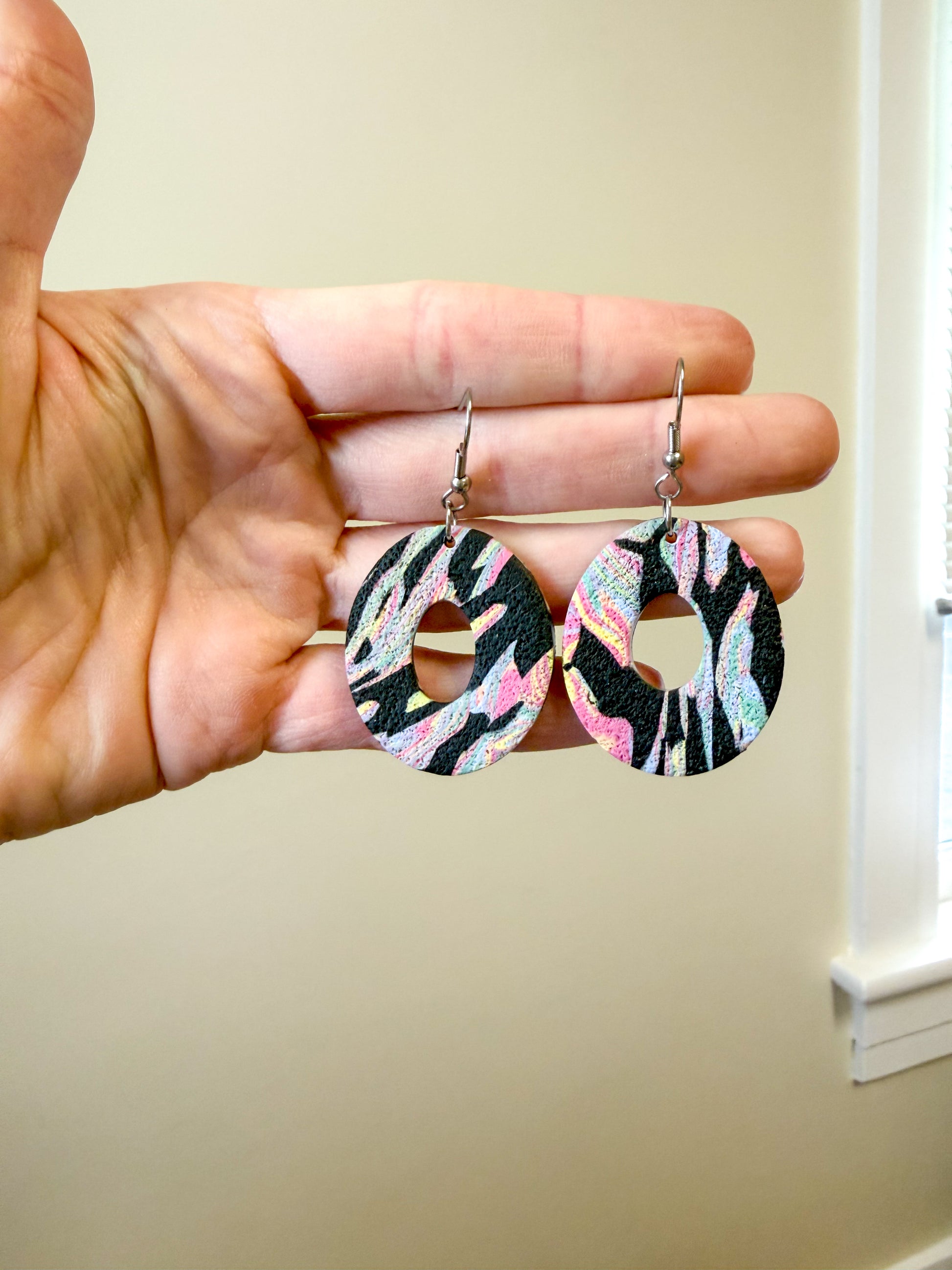 Colorful oval dangles with a center hole cut out. The pattern is black with colorful swirls. They are held in a hand 