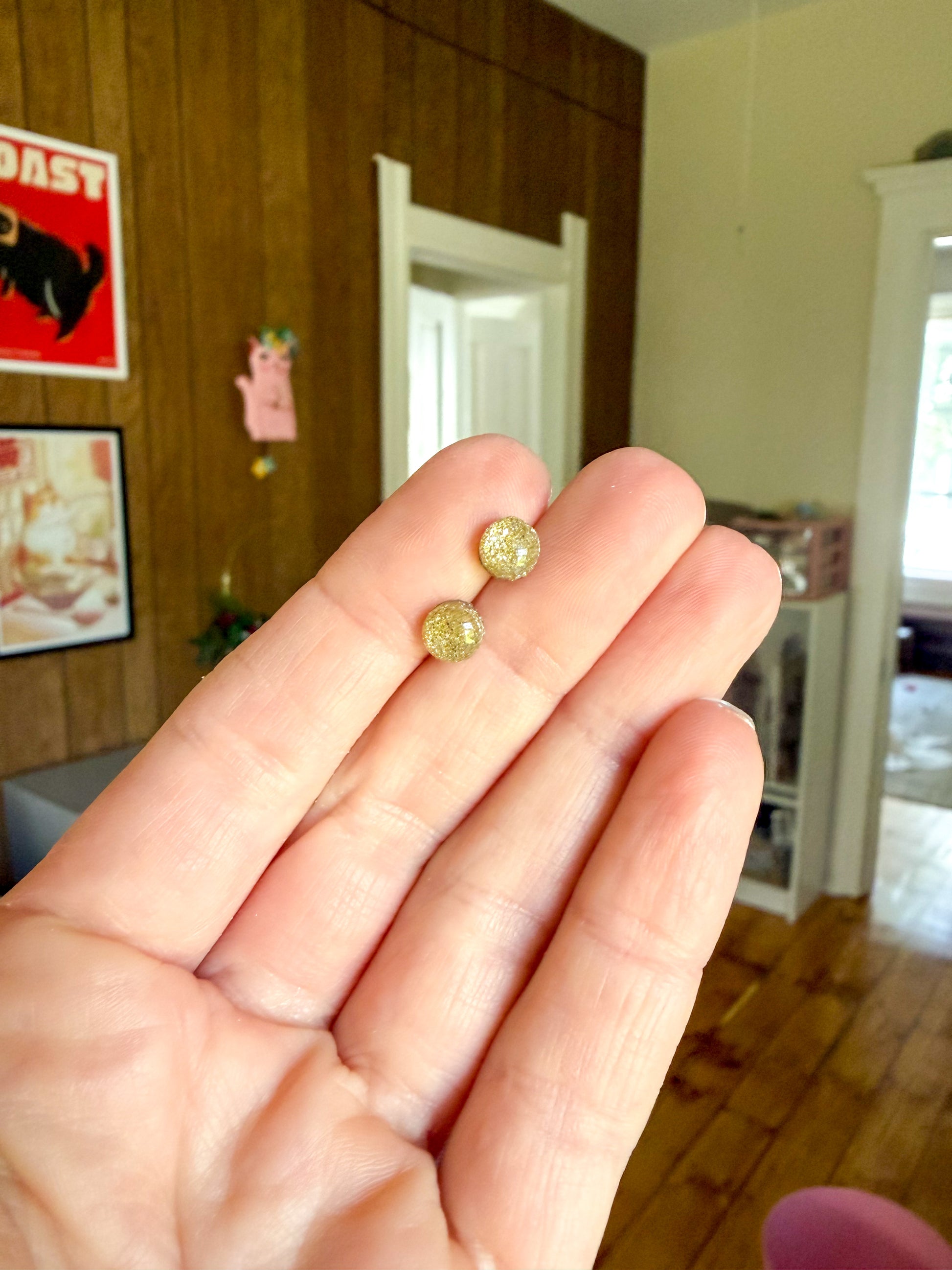 Two small round gold-toned earrings held on a person's fingers in an indoor room with wood-paneled walls and a wooden floor.
