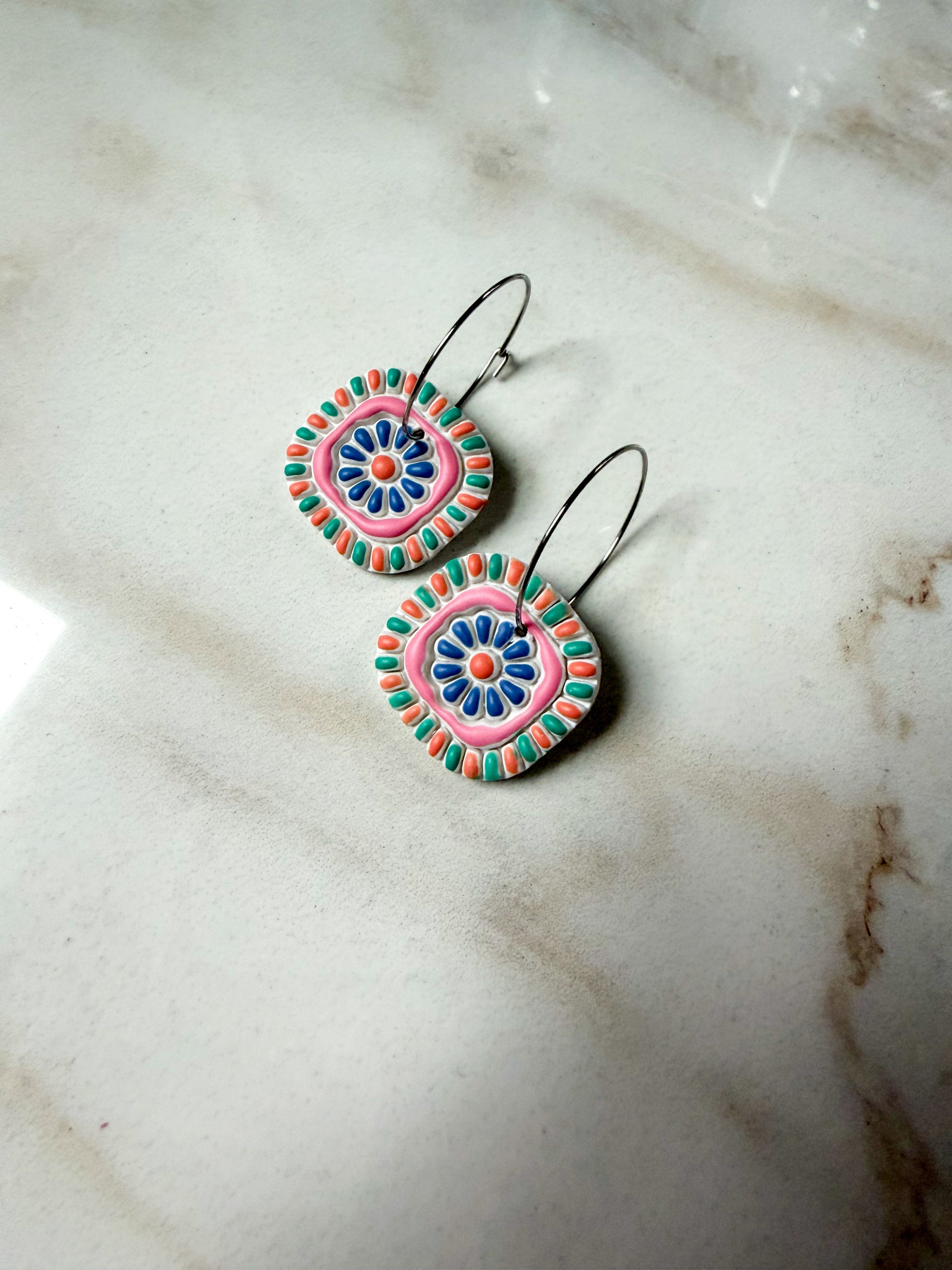 Colorful square earrings with silver colored hoops on a marble surface. 