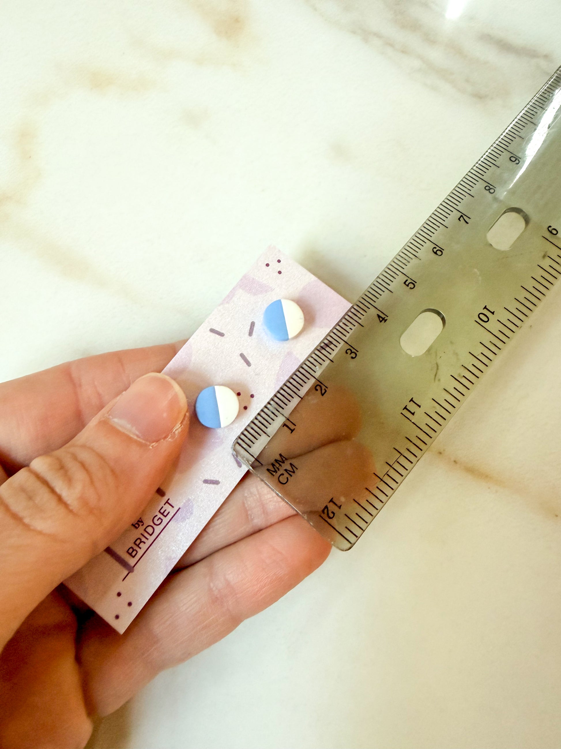 A hand holds a small purple card with two round stud earrings, alongside a clear plastic ruler, on a light marble-like surface. The card shows the brand name.