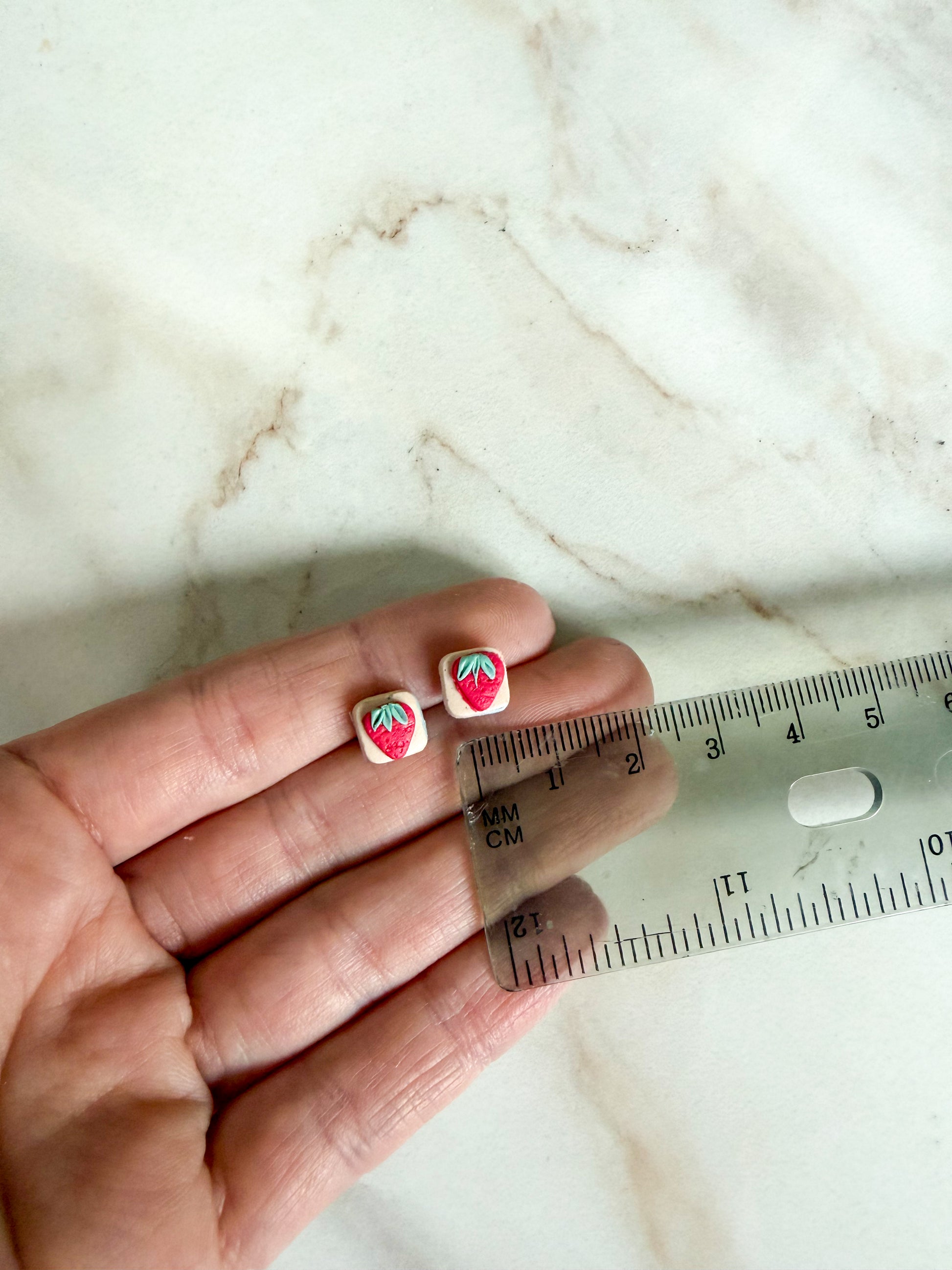 Small strawberry-themed earrings held between fingers with a ruler for scale on a marble surface. About 3/4 cm in length 