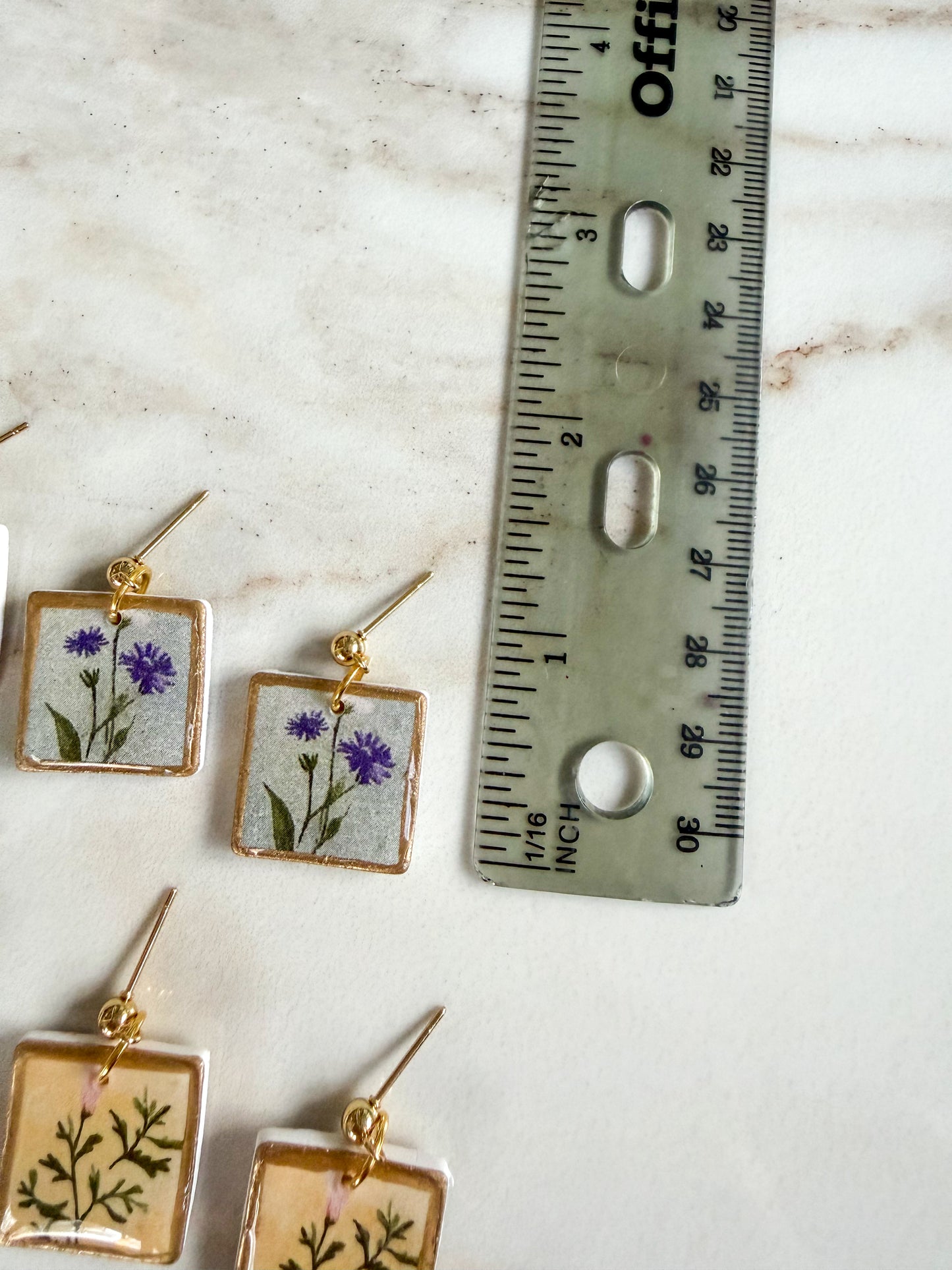 Four square earrings with floral designs on a marble surface next to a ruler. About 3/4 inches long 
