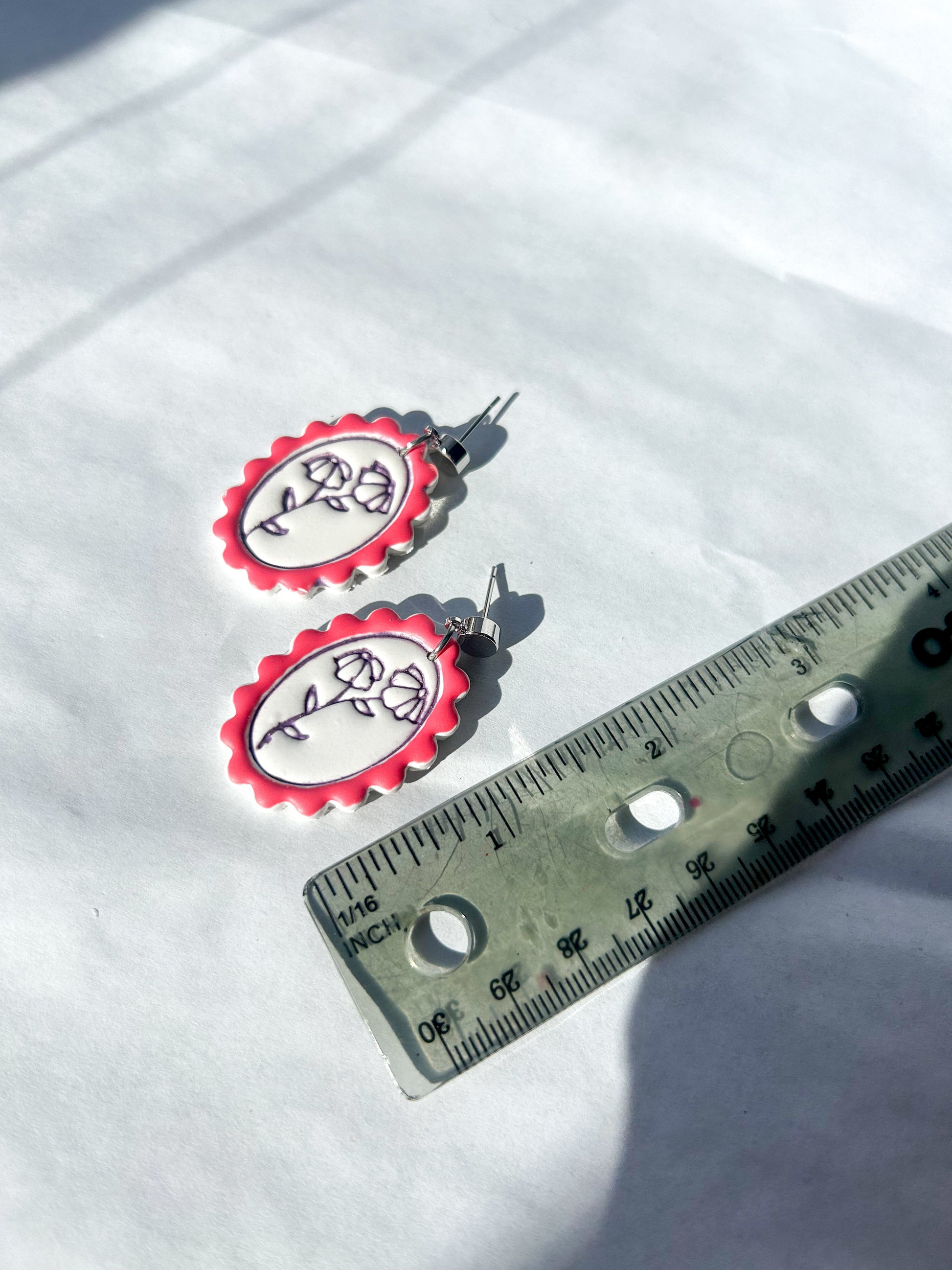Pair of earrings with red and white design on a white surface next to a ruler for scale.