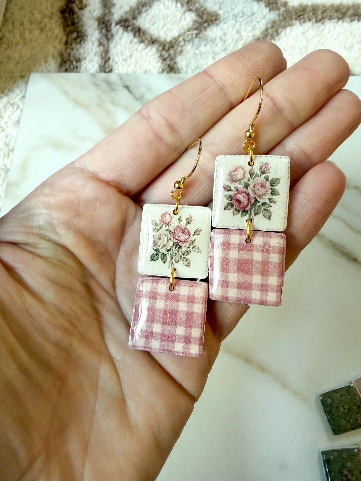 A pair of handmade dangle earrings held in a hand. The earrings feature gold fishhook wires, square white ceramic pinks with a pink rose floral print on the top and a pink and white gingham pattern on the bottom.