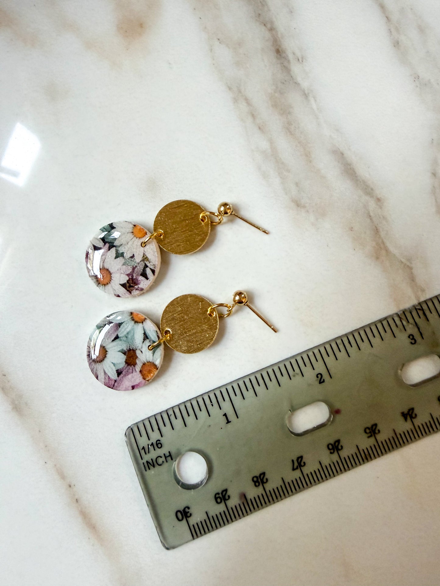A pair of double-drop earrings with one round floral resin charm and one textured gold disc charm, displayed next to a ruler on a white marble surface. About 2 inches in length. 