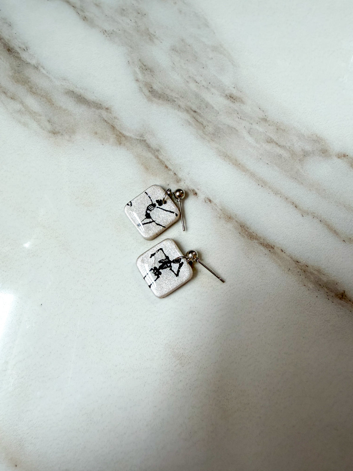A pair of small square earrings with black skeleton designs and silver fishhook backings, resting on a white marble surface.