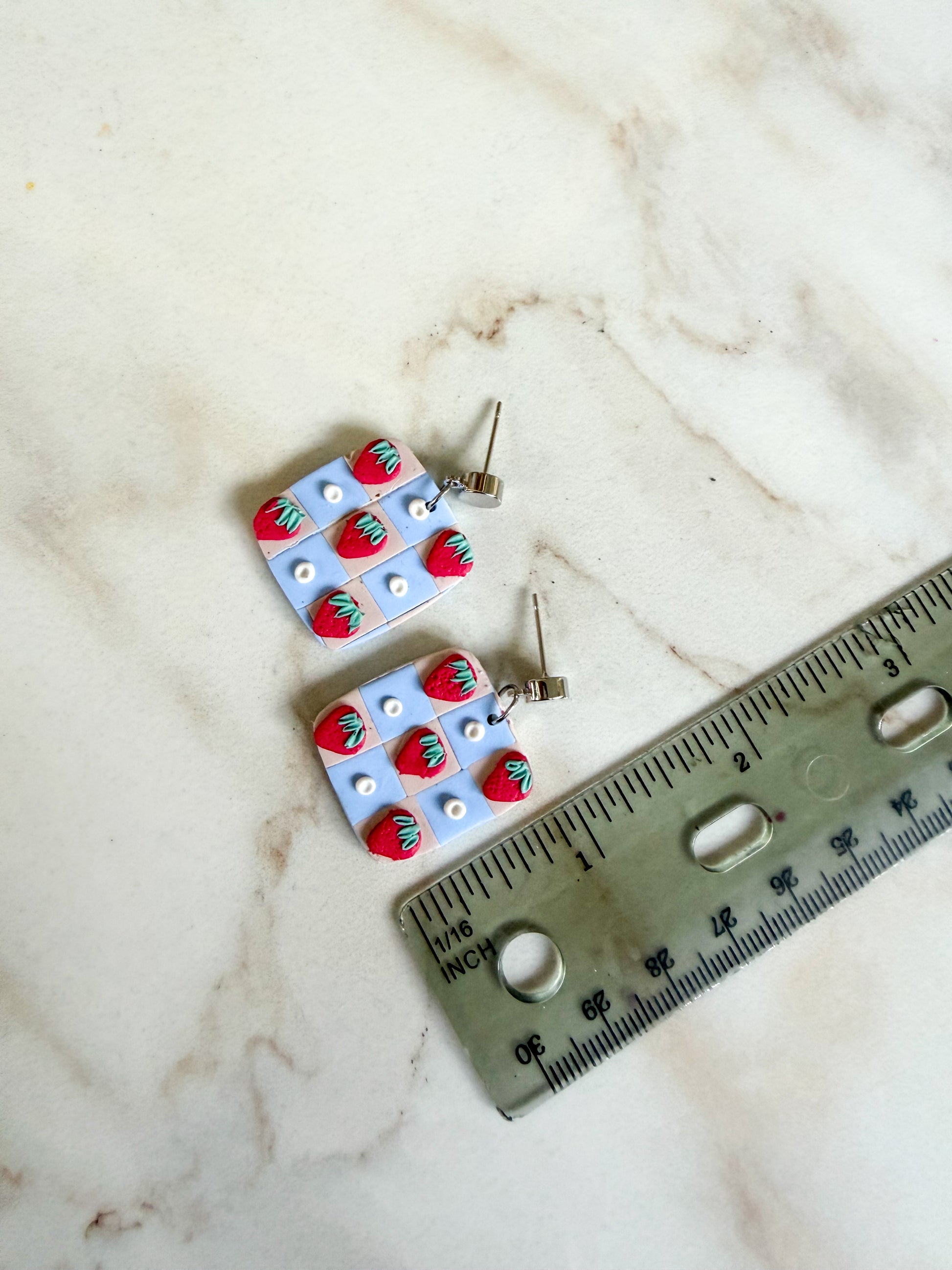 Pair of earrings with strawberry design on a marble surface with a ruler for scale. A little over one inch in length. 