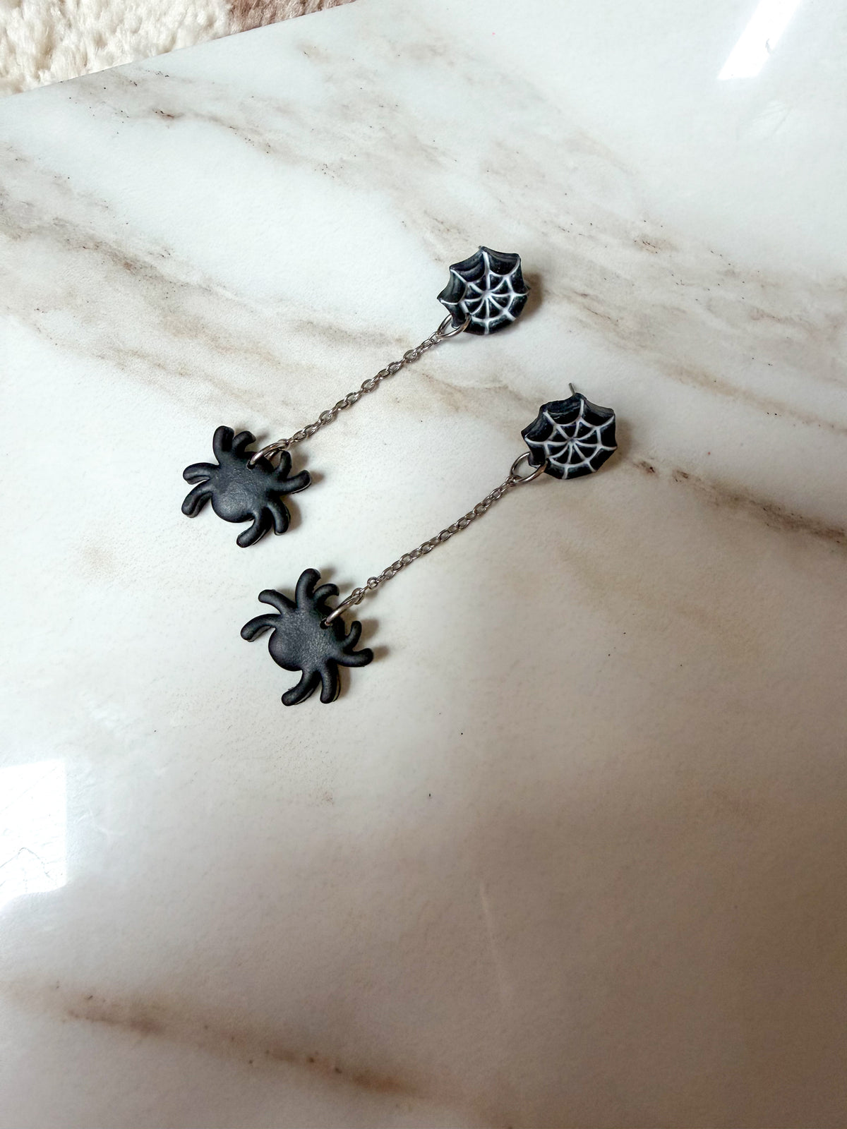 A pair of gothic spider drop earrings with black spiders and spiderweb charms on a silver chain, displayed on a beige marble surface.