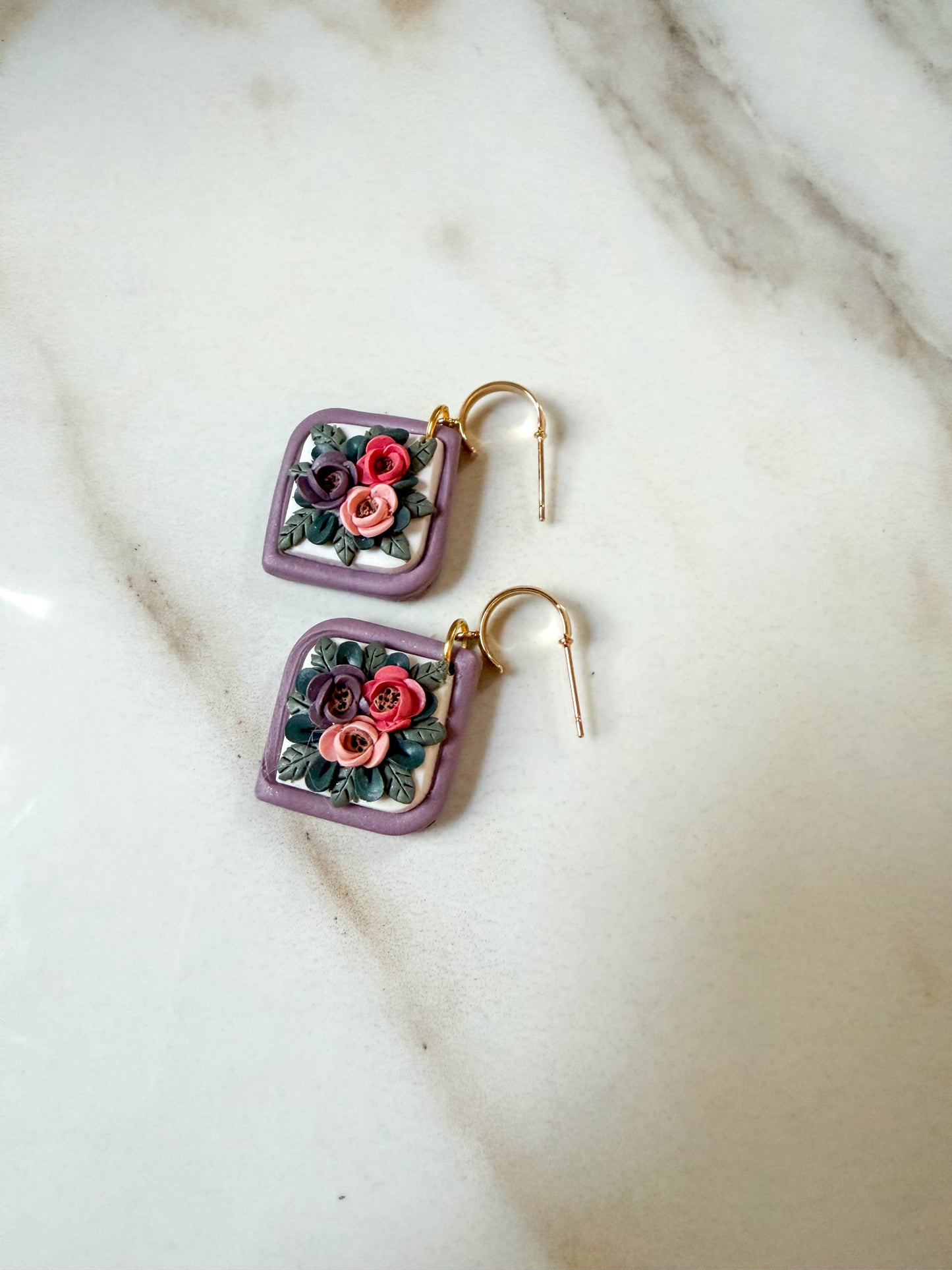 A pair of square lavender earrings with gold hooks and 3D clay floral designs in pink, red, and green, displayed on a white marble surface.