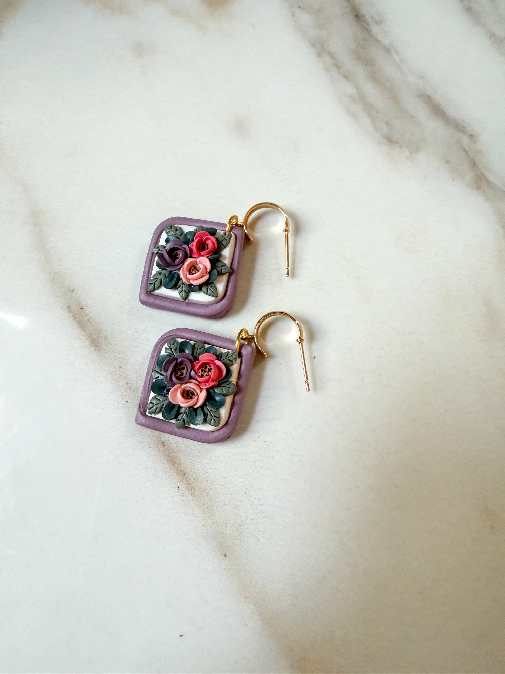 A pair of square lavender earrings with gold hooks and 3D clay floral designs in pink, red, and green, displayed on a white marble surface.