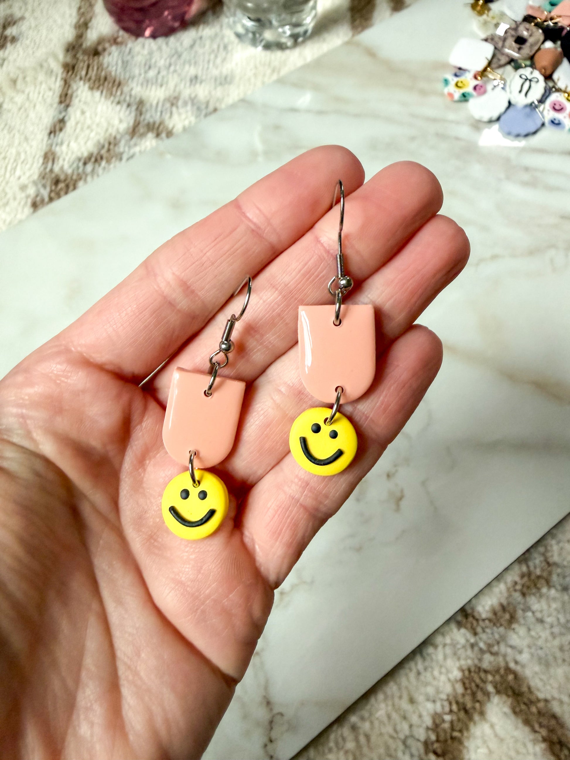 Hand holding a pair of earrings with smiley face charms on a marble surface.