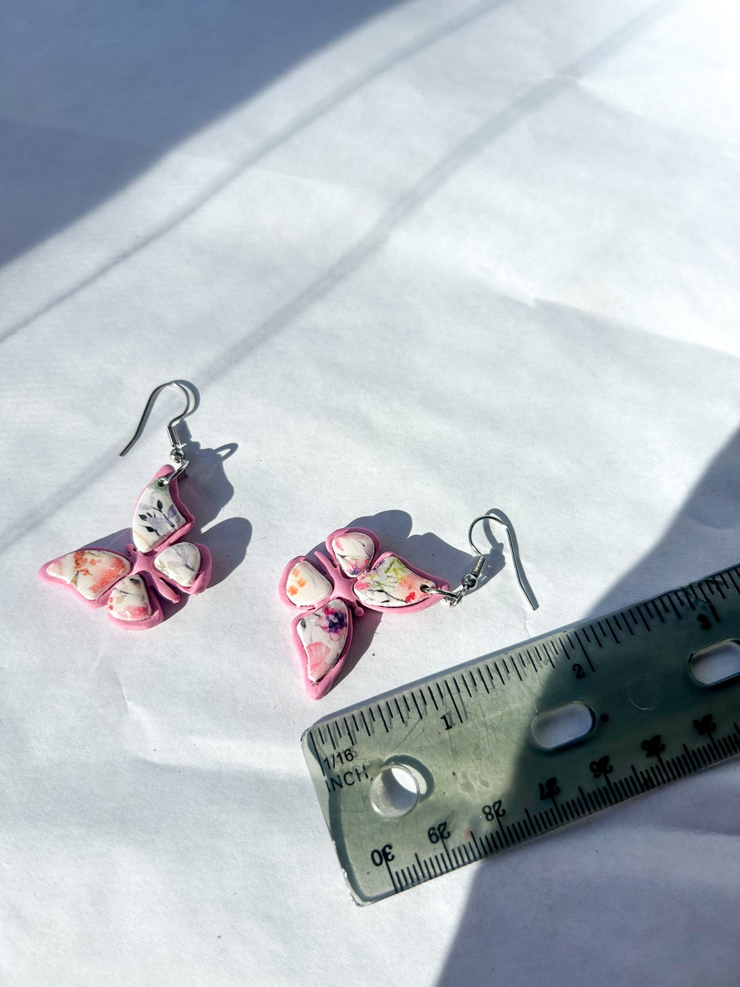 Pair of pink butterfly-shaped earrings on a white surface with a ruler for scale. About 2 inches in length. 