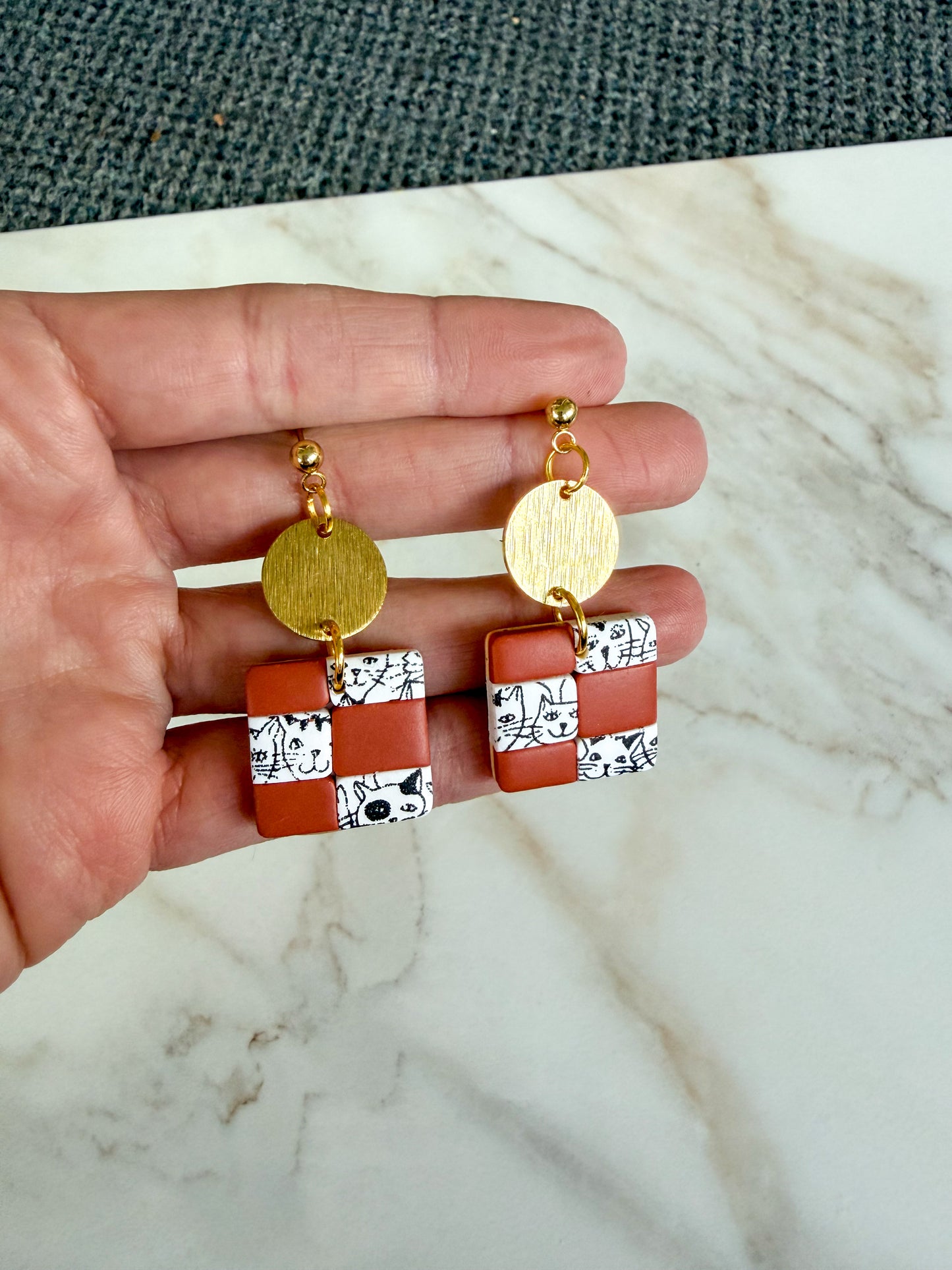 Gold and brown checkered earrings held in a hand on a marble surface