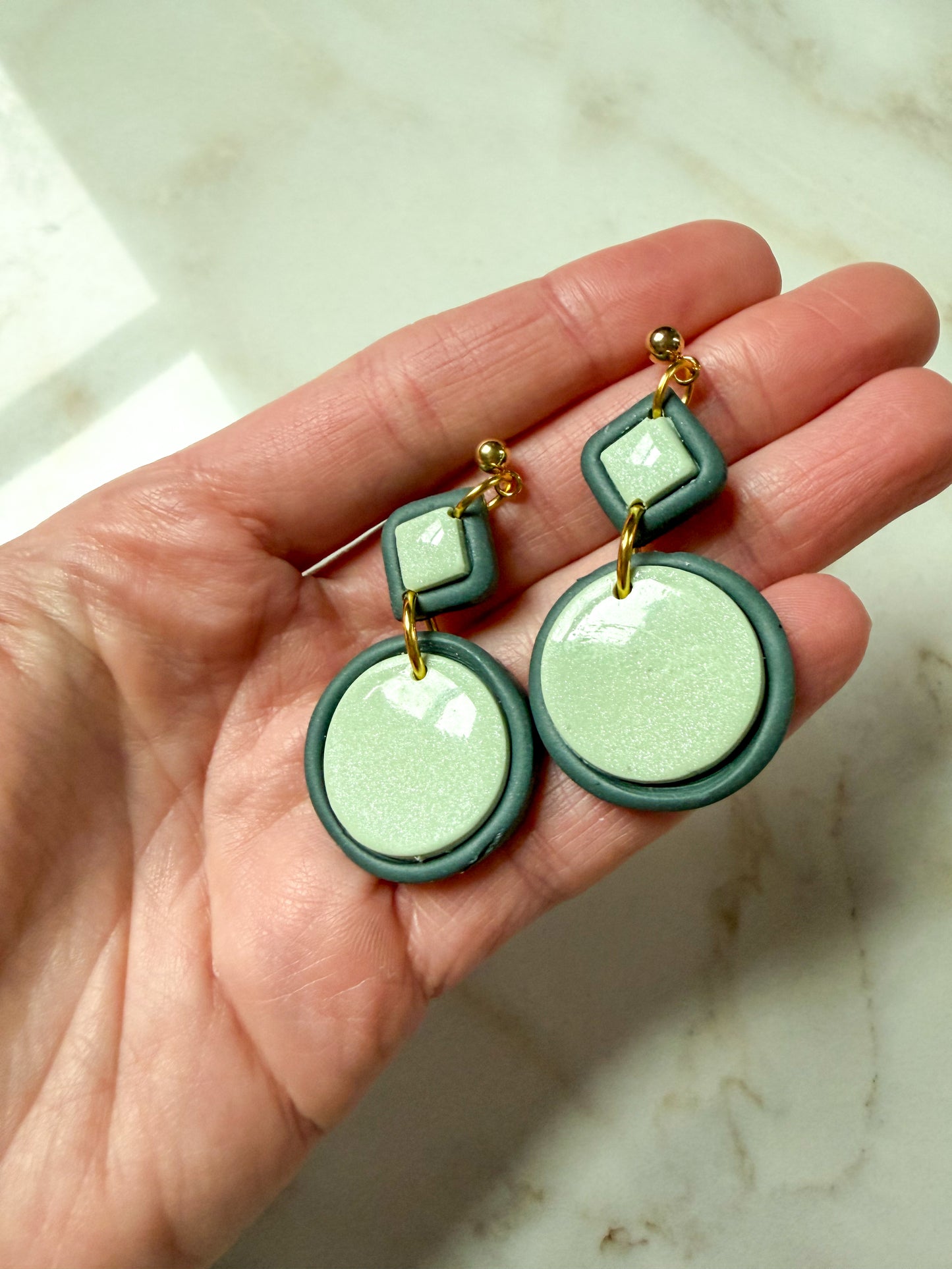 Green and gold earrings held in a hand on a marble surface