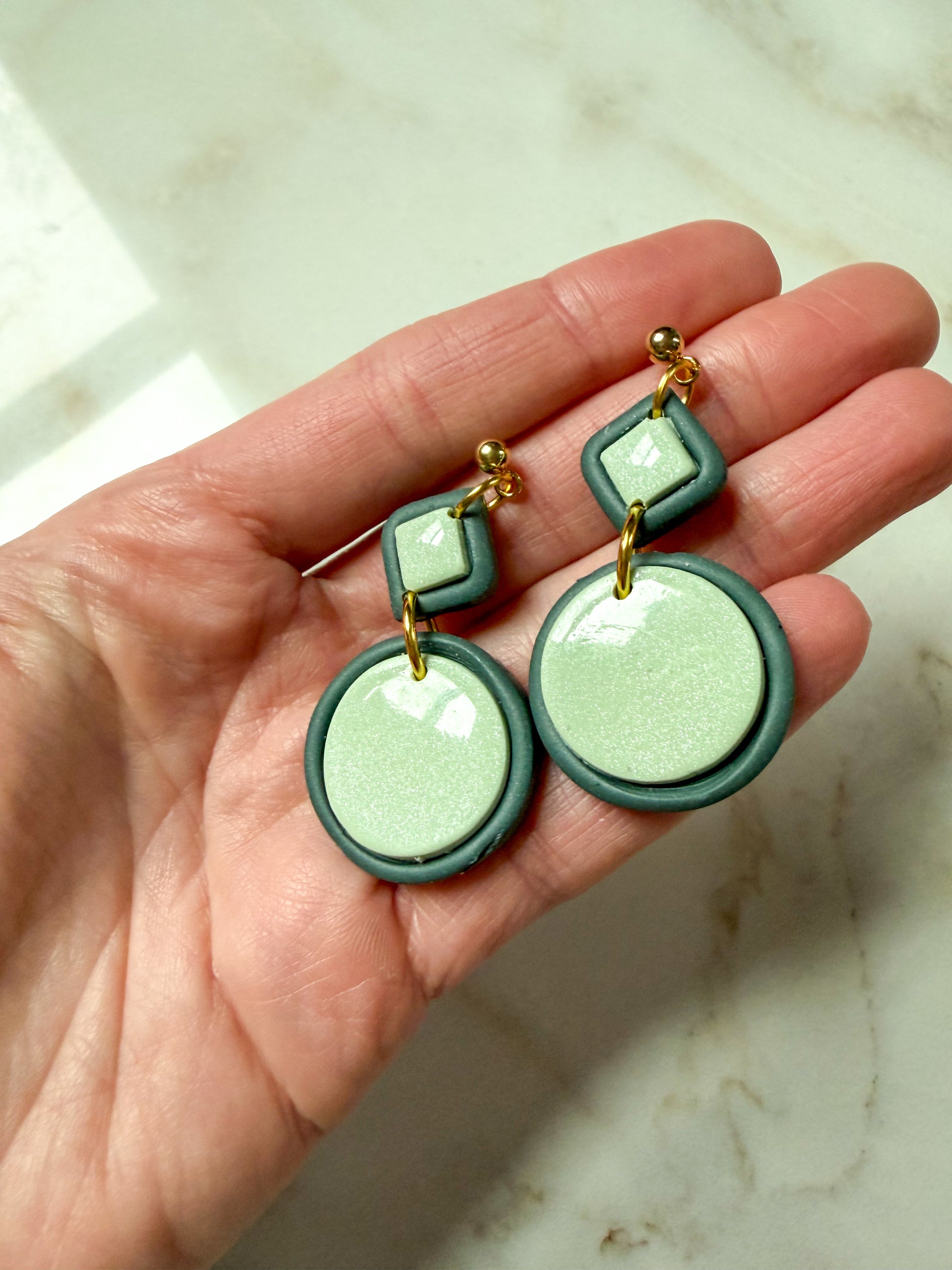 Green and gold earrings held in a hand on a marble surface
