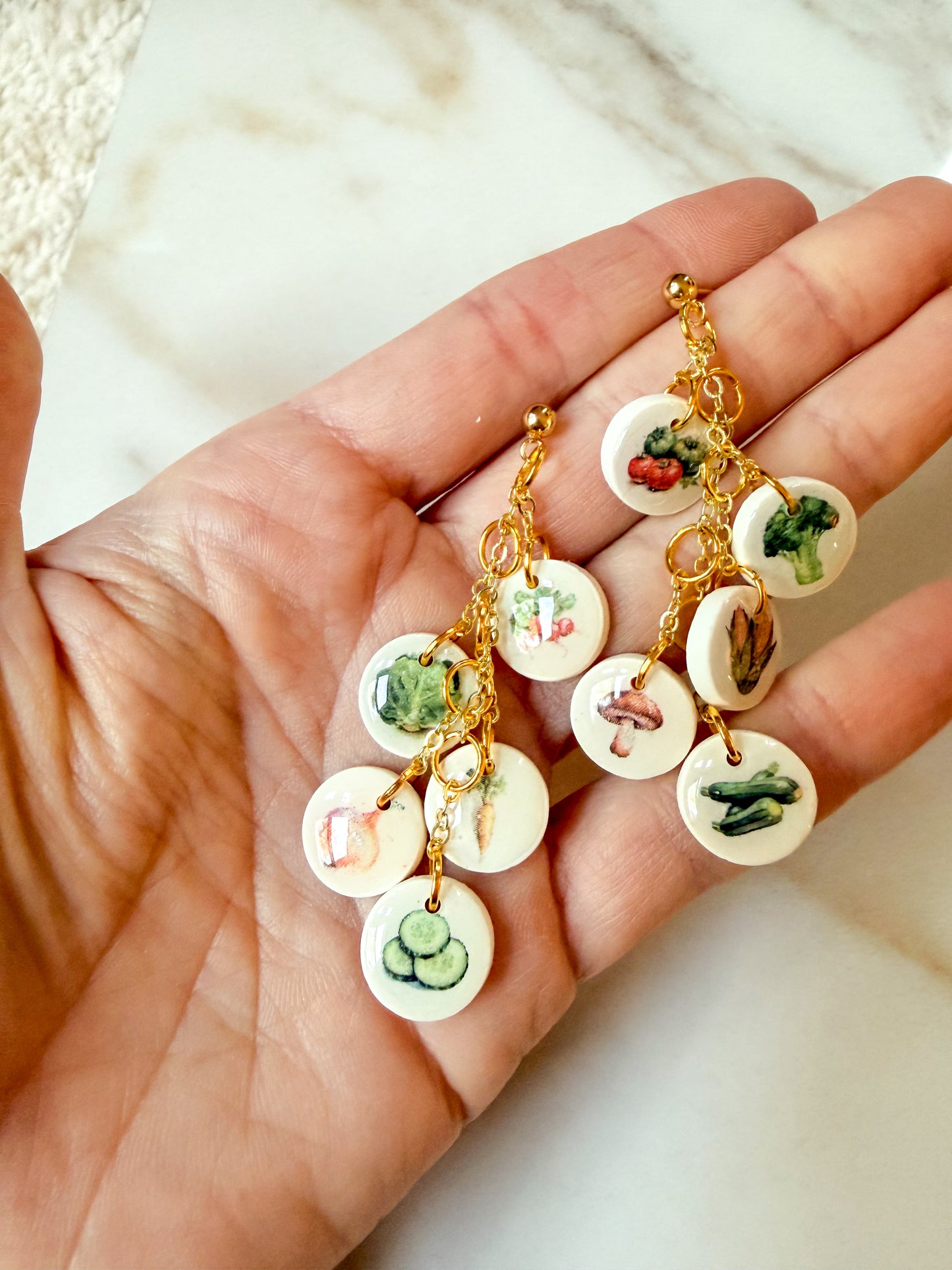 A pair of gold-tone dangle earrings with small round white charms printed with colorful vegetable illustrations, held in a person's hand against a light marble background.