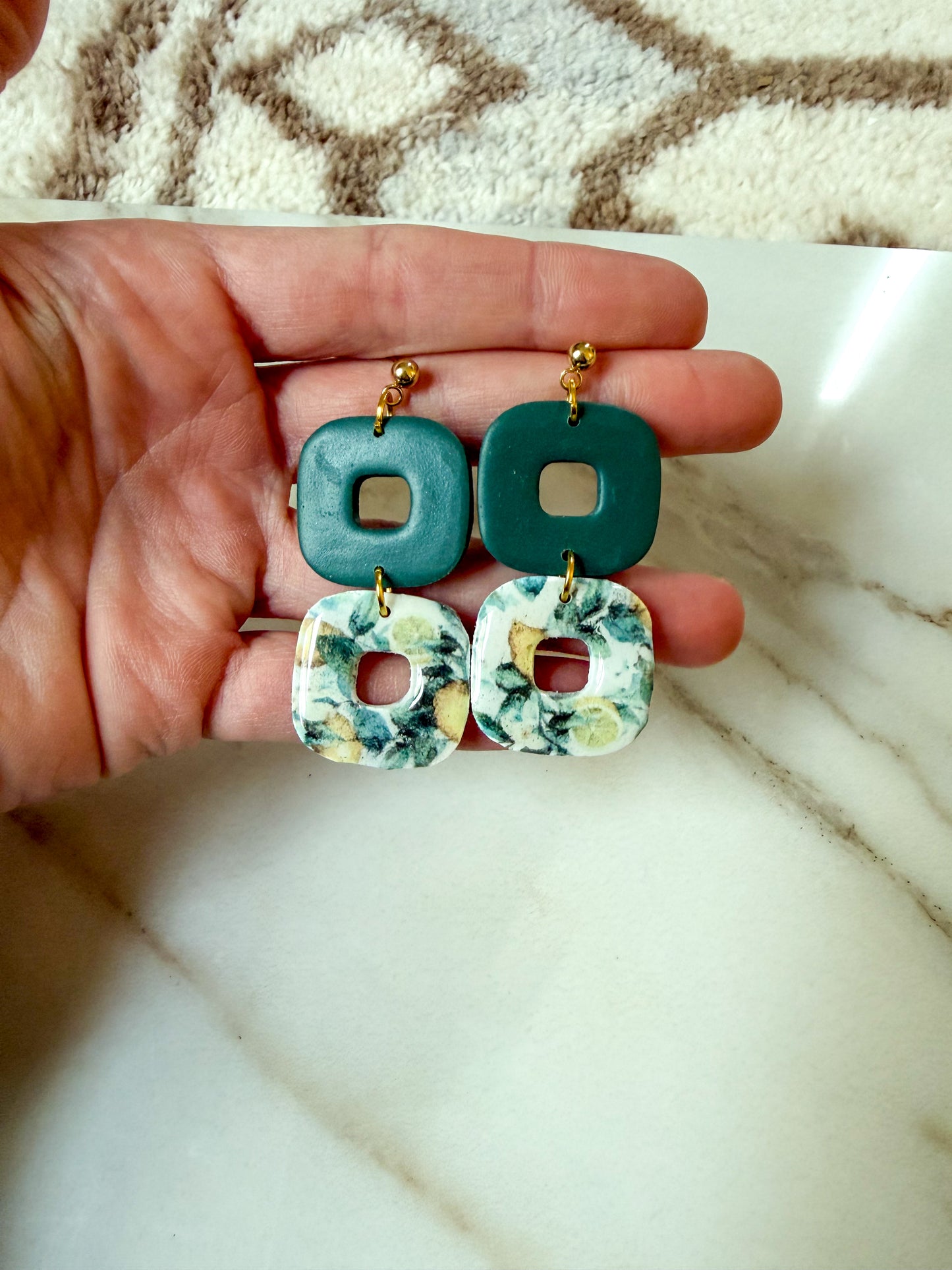 A pair of handmade square drop earrings held in a person's hand. The earrings feature a top dark teal green square with a square cutout, and a bottom white square with a green and yellow lemon pattern and a square cutout.