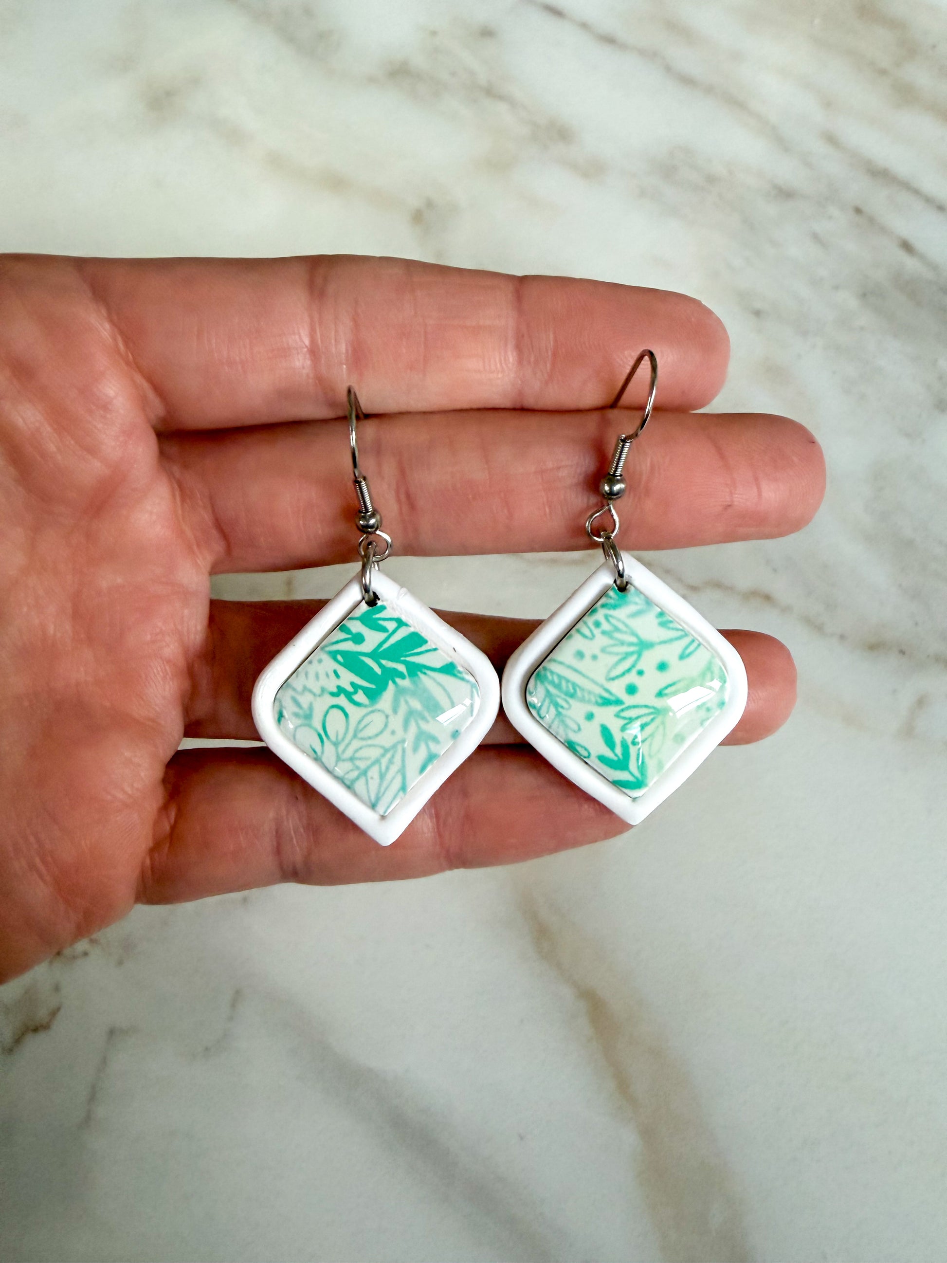 Pair of square earrings with green pattern held between fingers on a marble surface