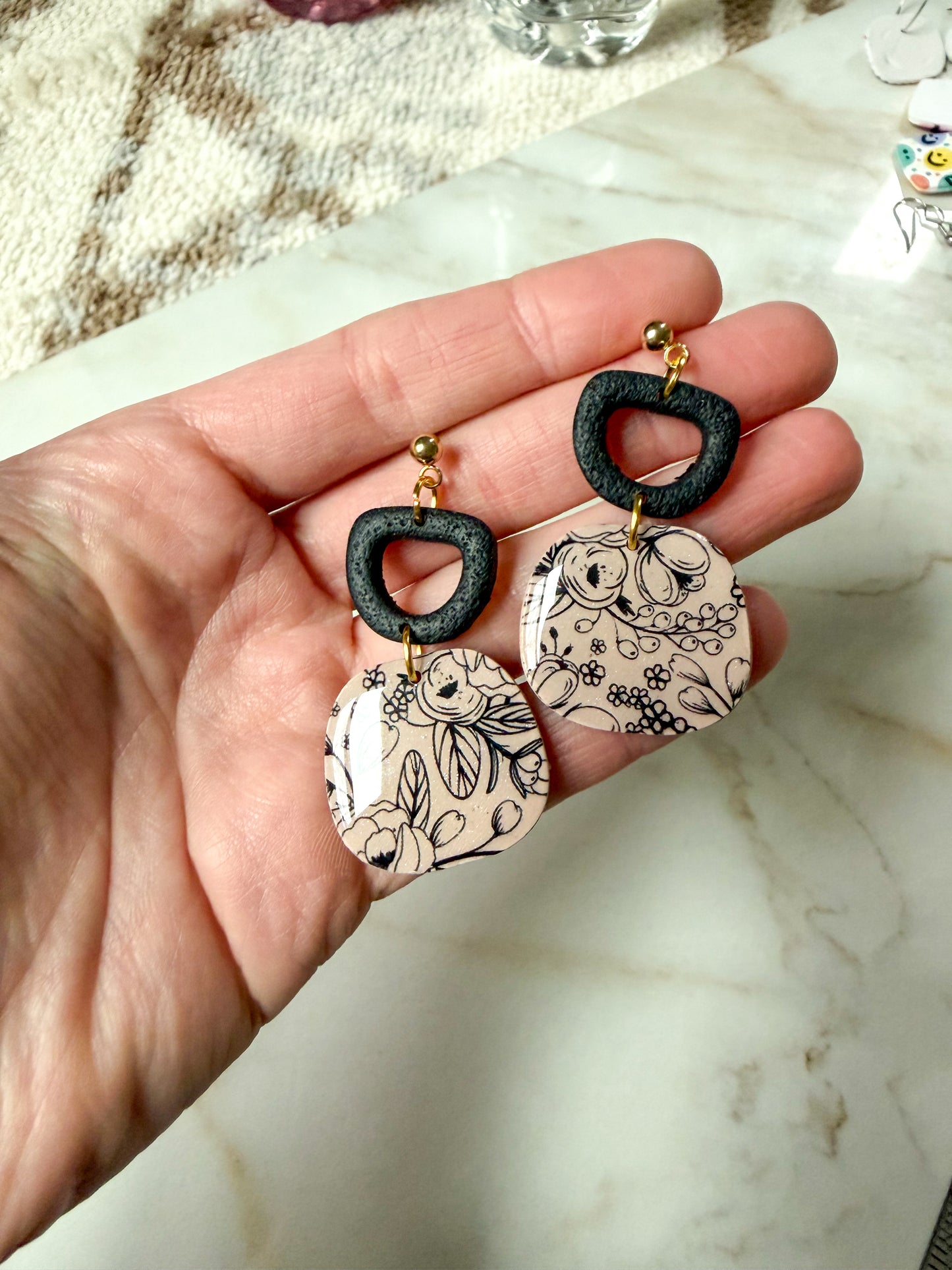 Hand holding a pair of earrings with floral designs on a marble surface
