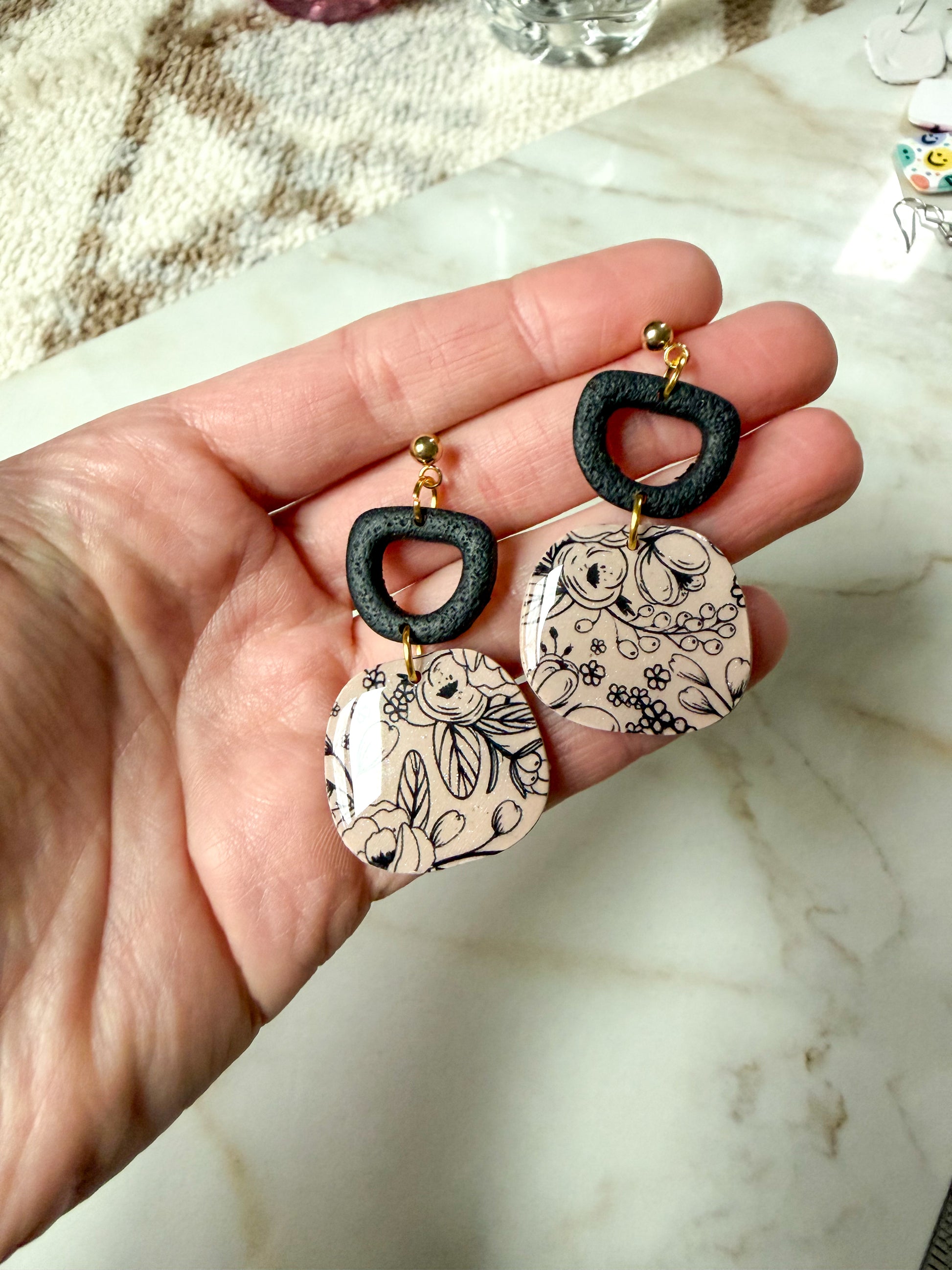 Hand holding a pair of earrings with floral designs on a marble surface
