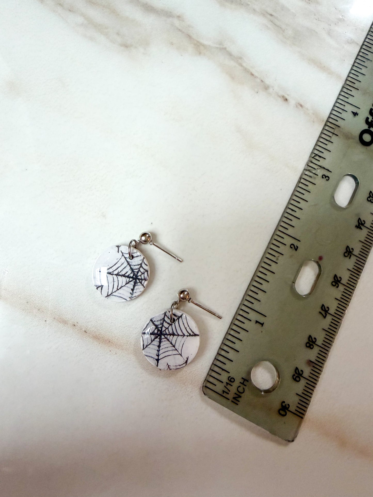 A pair of small round drop earrings with black spiderweb designs on a white background, shown next to a ruler on a light marble surface. About one inch long 