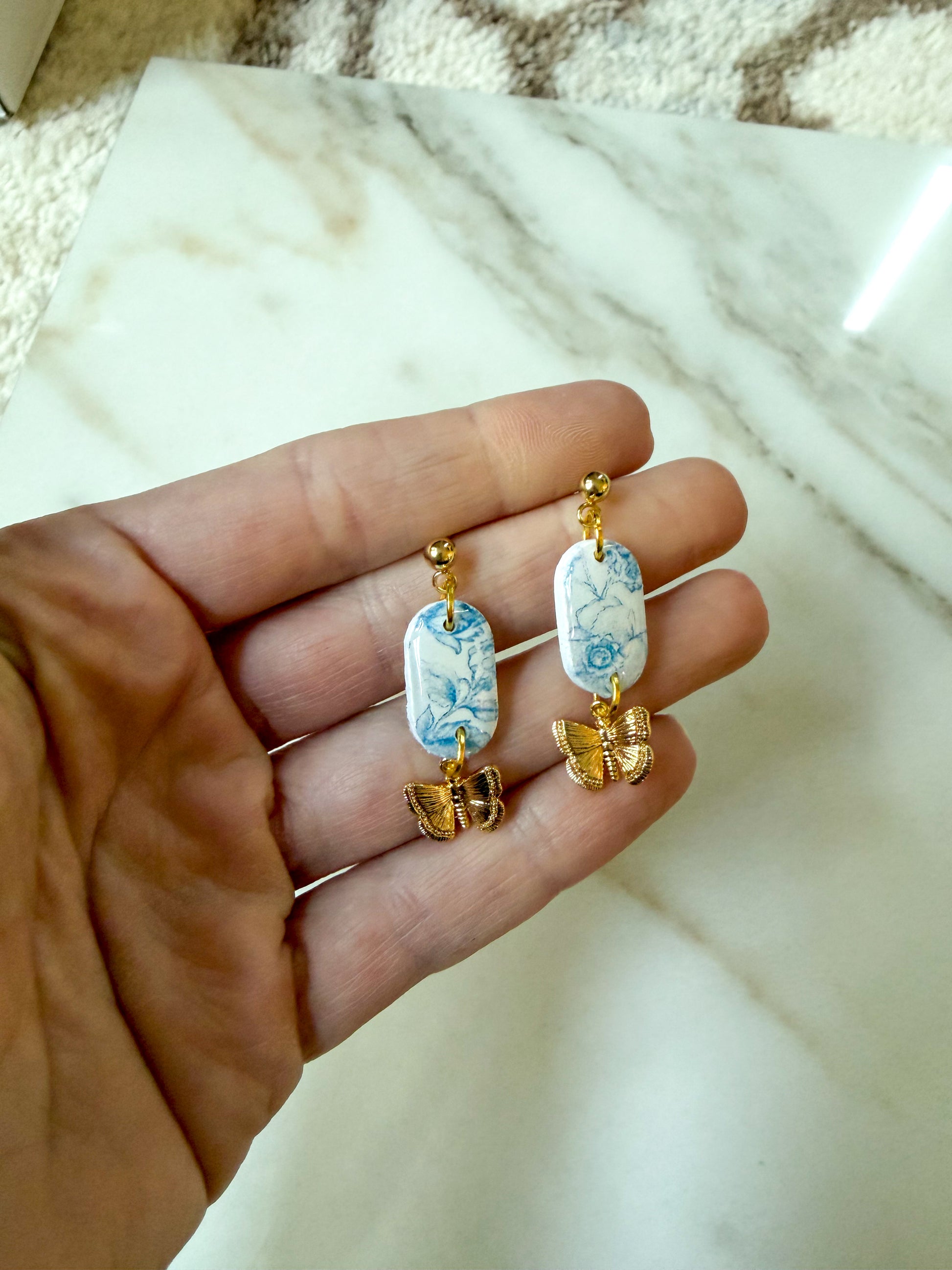 A pair of dangling earrings held in a hand, featuring white oval charms with a blue floral pattern and small gold butterfly charms below, set against a marble surface background.