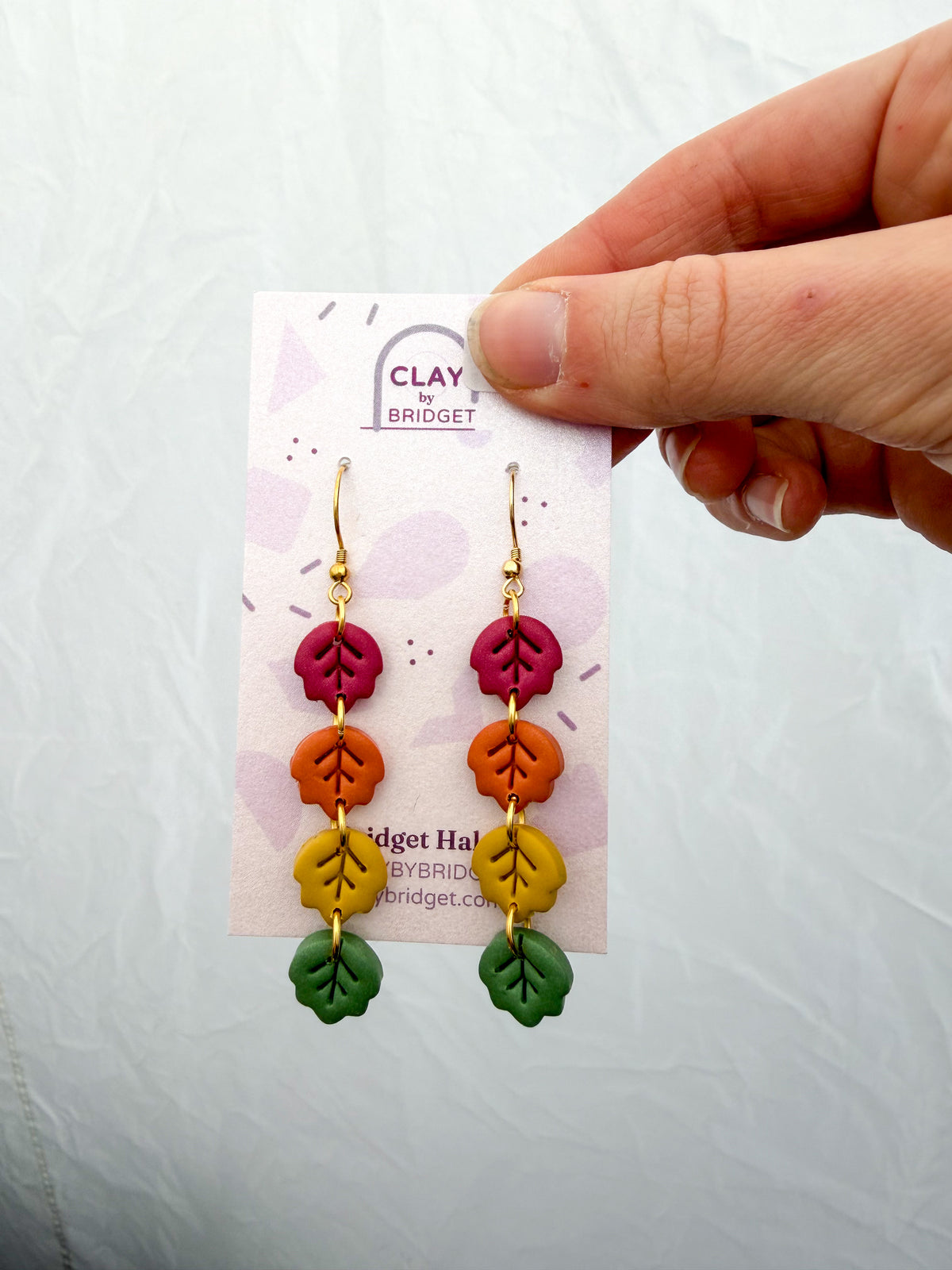A hand holds a pair of colorful leaf drop earrings on a display card. The earrings feature gold hooks with stacked leaf charms in red, orange, yellow, and green, shown against a white background.